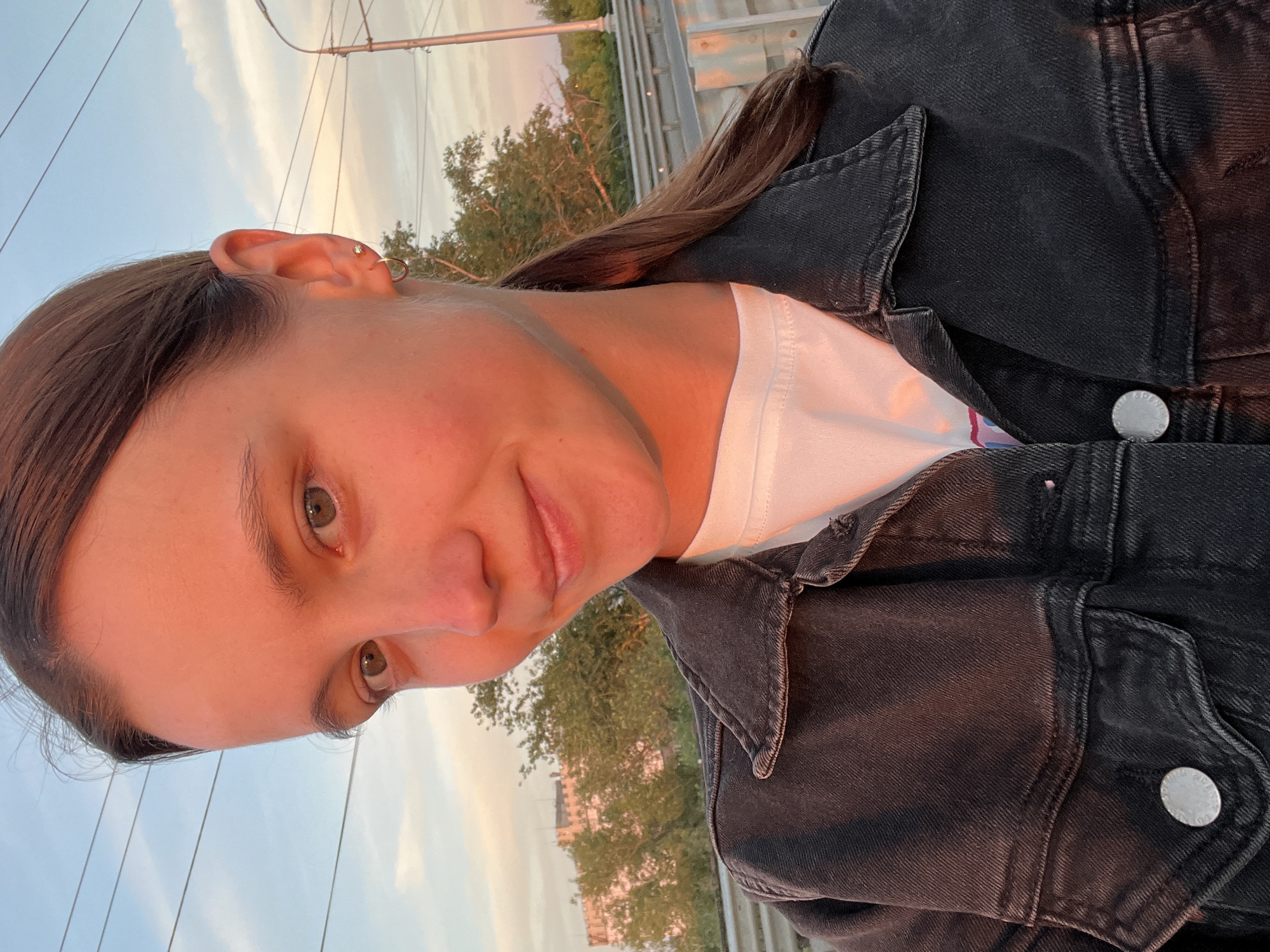 This is a self-portrait of a young woman taken outdoors. She is facing the camera with a slight smile and her head is tilted. She has long brown hair, pulled back, and is wearing a white t-shirt and a black denim jacket. She has a small stud earring in her upper ear and a hoop earring in her lower earlobe. The background shows a cloudy sky with hints of sunset colors, suggesting it is late afternoon or early evening. Trees and a railing are visible behind her, and some power lines stretch across the sky. The image appears to be taken in Novokuybyshevsk, Russia. There is no visible text in the image.