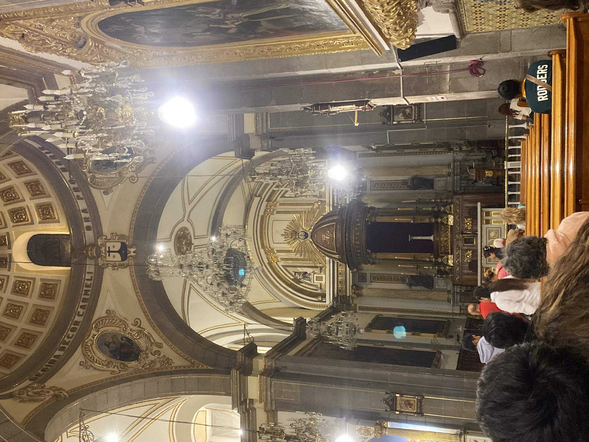 The image captures the interior of the Templo del Carmen in Puebla, Mexico. The shot is taken from the perspective of someone seated in a pew, facing the altar. Elaborate chandeliers hang from the intricately decorated ceiling. People are seated in the pews, with their backs to the viewer. The altar, adorned with religious iconography and candles, is the focal point. The architectural details, including arches, pillars, and gold accents, are prominent. The lighting is a mix of natural and artificial, casting shadows and highlighting certain areas.