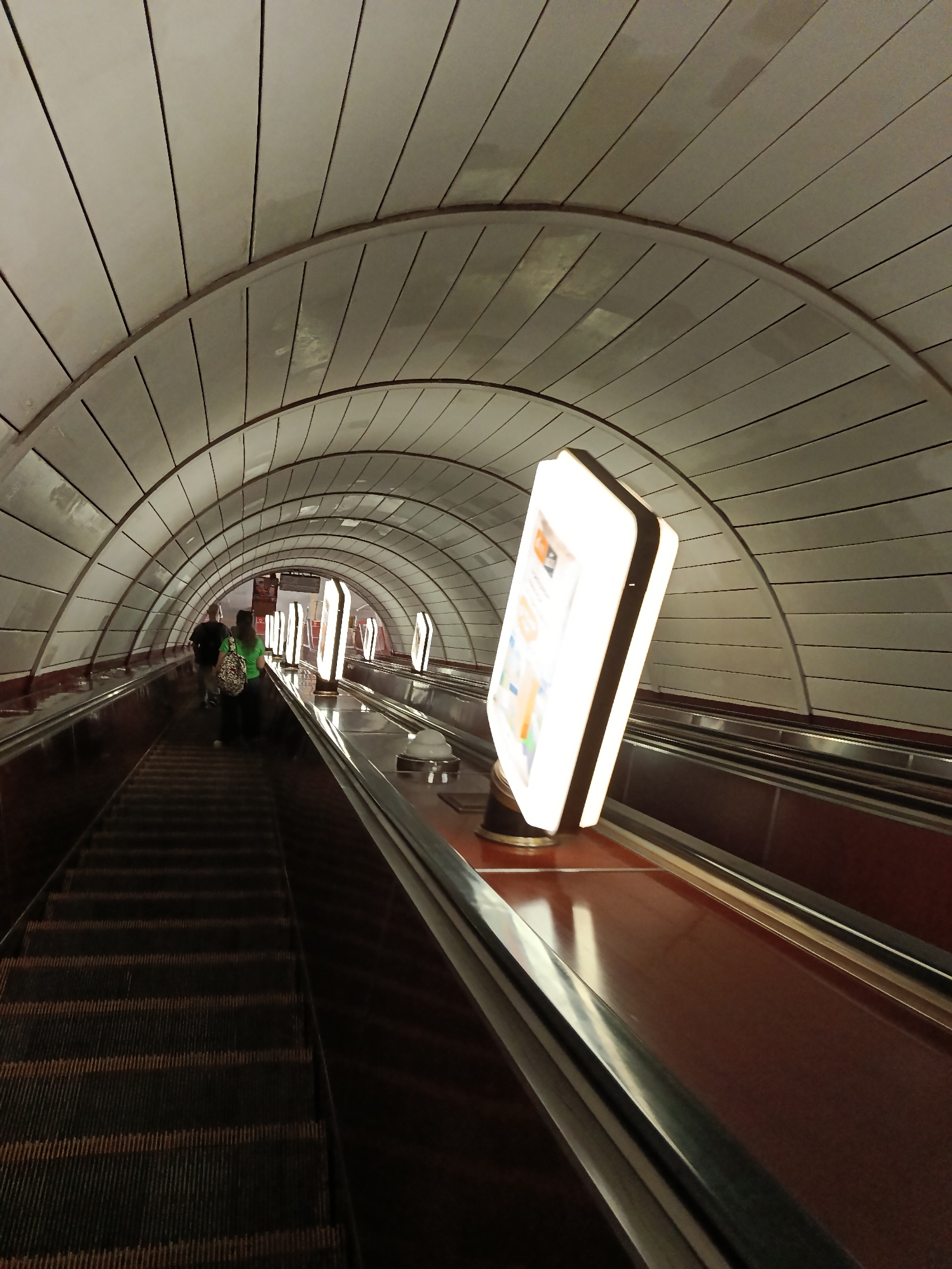 The image depicts a subway escalator tunnel with two people descending. The tunnel features a distinctive arched ceiling constructed of white panels with visible seams and metal support beams. The escalators themselves have dark treads and reddish-brown sides. Several illuminated signs are mounted on posts along the escalators, one of which is prominently in the foreground, displaying colorful graphics and text. Two individuals are visible in the background, walking down the escalator. The overall impression is one of an underground public transportation environment, likely a subway station. The lighting is artificial and bright, illuminating the tunnel and the signs. There is no indication of the time of day or weather. The mood is neutral, reflecting a typical urban transit scene. The visible text on the sign in the foreground is illegible.
