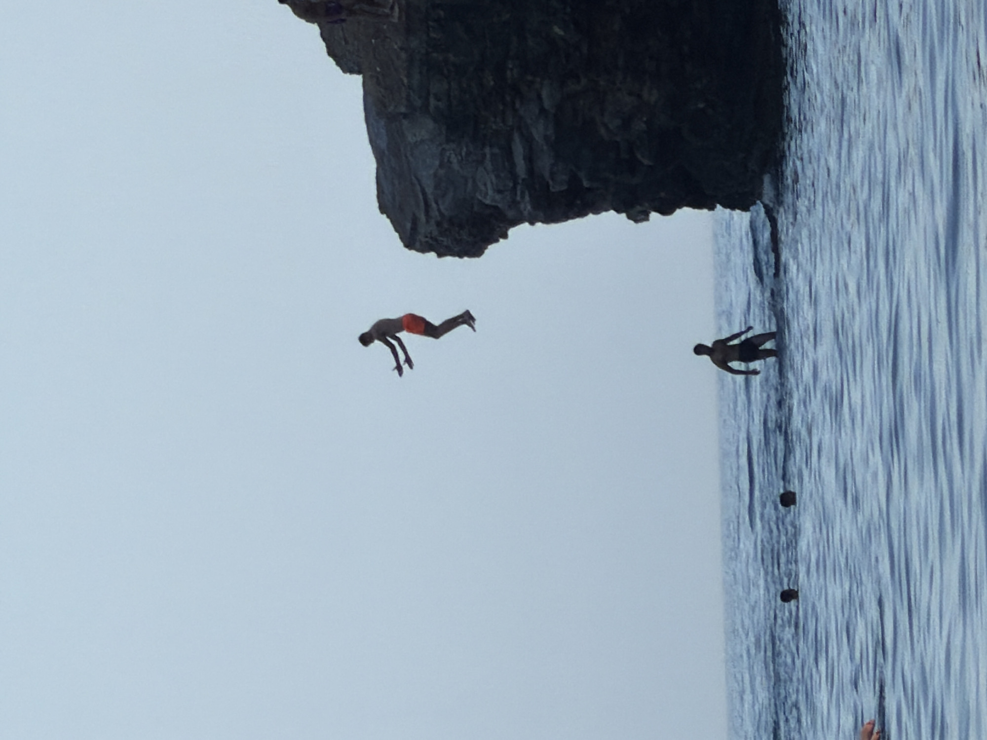 The image depicts a group of people engaged in recreational activities by the sea, with a prominent rock formation serving as a backdrop. In the foreground, the deep blue, choppy water suggests a moderately active sea. Several individuals are visible in the water, with two heads just breaking the surface and another person wading near the shore.
The central focus of the image is a person in mid-air, having jumped from a high rocky outcrop. This individual is wearing orange shorts and appears to be in the process of a dive or jump, with arms outstretched. The rock formation itself is rugged and dark, indicative of natural coastal erosion. Another person is standing on the edge of this same rock formation, also appearing to be preparing to jump or observe.
The sky is overcast with a pale, hazy blue, suggesting either early morning, late afternoon, or a cloudy day. The overall scene is one of outdoor recreation and adventure.
While the image depicts a scene consistent with coastal areas worldwide, without specific landmarks or signage, it is impossible to definitively pinpoint its exact location within Budva, Montenegro. Budva is known for its beaches and rocky coastline, so such an activity is plausible there. However, a precise identification requires more contextual information not present in the visual data alone.