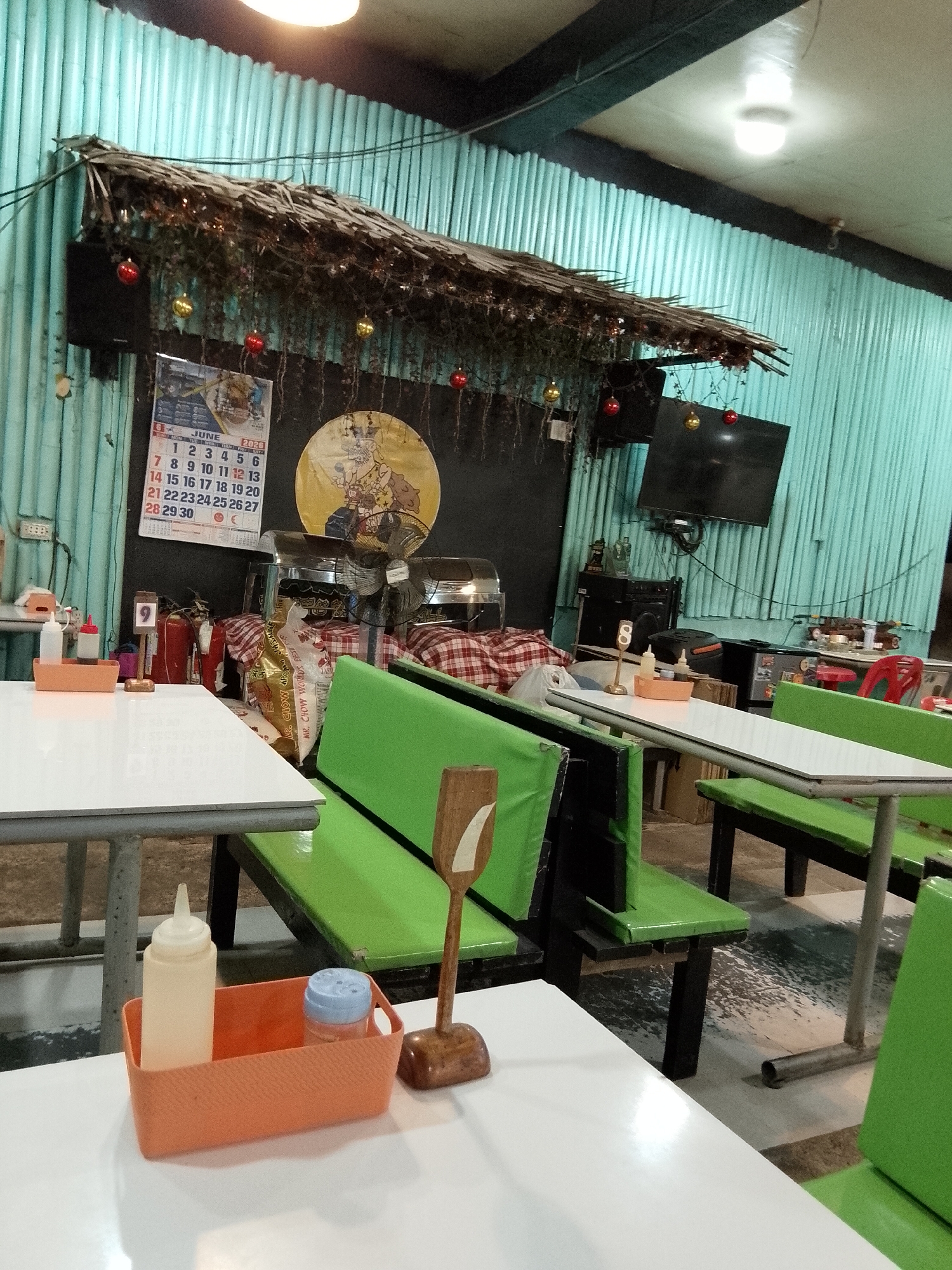 The image depicts the interior of what appears to be a casual dining establishment or eatery, possibly a carinderia, in Davao City, Philippines. The setting is rustic, with walls made of corrugated metal painted a light turquoise. A thatched roof structure, adorned with Christmas ornaments (red and gold balls) and hanging vines, is visible above.
In the foreground, a white table with a metal frame is set with an orange condiment caddy containing a squeeze bottle and a shaker. A wooden utensil holder with a number "7" is also present on the table. To the right, a bright green bench with a black wooden frame is partially visible. Another table and bench are visible in the background, also with green seating.
The middle ground of the image reveals a variety of objects. A calendar for June 2020 is hanging on the wall, displaying a grid of dates. Next to it is a round decoration featuring a cartoon character. In front of the wall, there are food warming trays, a fire extinguisher, and stacks of checkered fabric. A large bag of what looks like snacks or ingredients is placed on a bench. On the right side of the image, a flat-screen television is mounted on the wall, and below it is a stereo system. Further to the right, more tables and seating can be seen, along with a red plastic chair. The floor is tiled and appears slightly wet or stained in some areas.
The lighting in the scene is a mix of overhead fluorescent lights and ambient light, creating a somewhat dim atmosphere. The overall impression is of a simple, functional eatery, likely in a local neighborhood of Davao City.