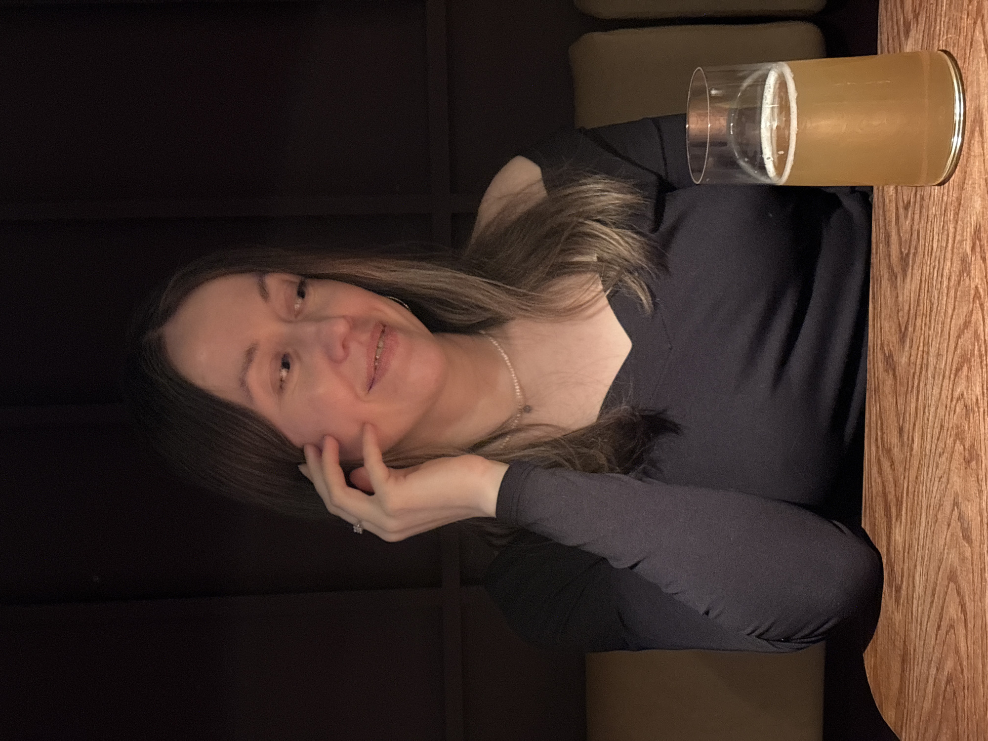 A woman sits at a wooden table indoors. A glass of beer sits on the table beside her. The location is impossible to determine from the provided image.