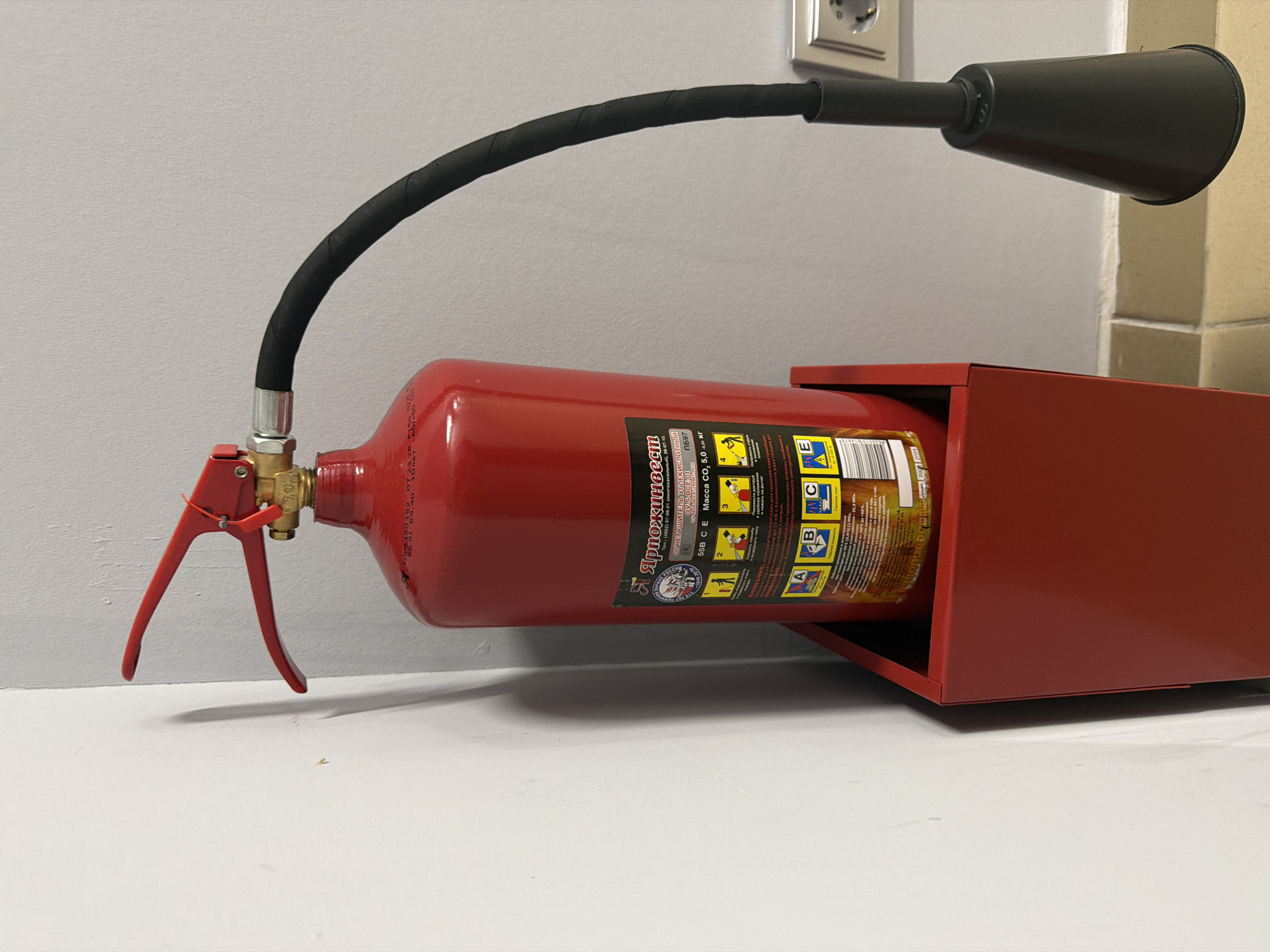 The image displays a red carbon dioxide fire extinguisher, mounted horizontally on a light-colored wall, characteristic of an indoor setting such as an office or public building in Saint Petersburg, Russia. The fire extinguisher is secured within a matching red metal holder or bracket.
A black, corrugated hose extends from the brass-colored valve assembly of the extinguisher, curving upwards and then horizontally to terminate in a black, horn-shaped nozzle. The extinguisher's trigger mechanism is red, with a red safety tie attached near the handle.
The label on the fire extinguisher, predominantly in Russian, provides detailed information. Key visible text includes the brand "Ярпожинвест" and the description "ОГНЕТУШИТЕЛЬ УГЛЕКИСЛОТНЫЙ" (Carbon Dioxide Fire Extinguisher). Further specifications read "ОУ 5 ВСЕ 01" and "55B CE Macca CO₂ 5.0 кг" (mass of CO₂ 5.0 kg). The label also features "ВНИМАНИЕ:" (Attention/Warning), a barcode, and illustrated instructions for use in four numbered steps, along with symbols for fire classes A, B, C, and E. Contact information, "Тел.: (4852) 67-96-01", is also printed.
The background consists of a textured, light-colored wall. A power outlet is visible on the wall in the upper right quadrant, alongside a cream-colored architectural element, possibly a pillar or corner. The fire extinguisher rests on a light, possibly white, flat surface below its mount. The scene shows the equipment in a state of readiness for emergency use.