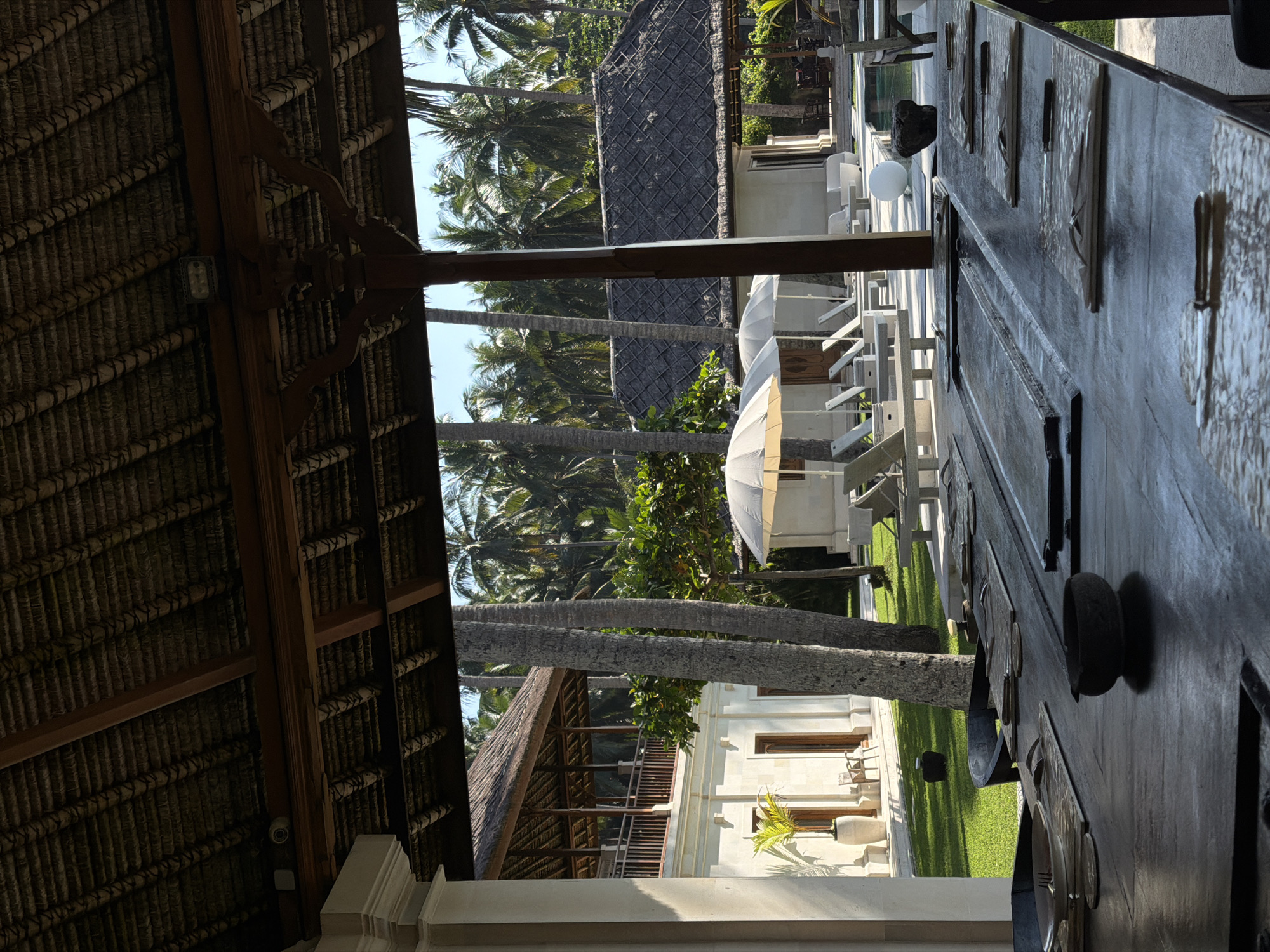 The image captures an outdoor dining setting in Karangasem, Indonesia. A long, dark wooden table set for a meal dominates the foreground. It is adorned with tableware. The view extends through the open-air structure towards a tropical landscape. Palm trees and white structures dot the scene, including a building with a thatched roof, and white lounge chairs with a white umbrella. Green lawns are visible. The perspective is from under a thatched roof structure, adding depth and framing the scene.