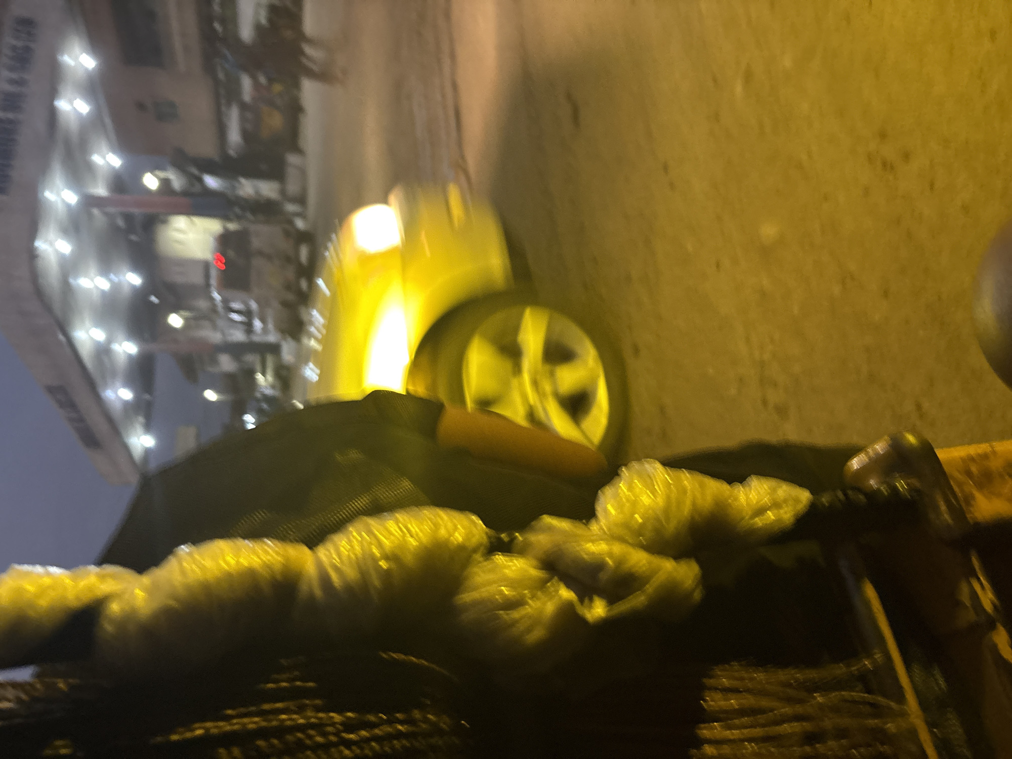 The image, taken at night in Onitsha, Nigeria, captures a street scene dominated by artificial lighting and a yellow-toned hue. In the foreground, a person's braided hair is visible on the left, with what appear to be items wrapped in yellow plastic bags. A dark object, possibly a vehicle or structure, is to the right of the person.
The middle ground features a bright yellow car, blurred due to motion or focus, with its headlights illuminated. The car is on a paved surface that appears to be a road. Behind the car, a structure with multiple lights is visible, suggesting a commercial establishment, possibly a gas station given the canopies and the presence of vehicles. The lighting suggests it's an open-air area.
In the background, the sky is dark, indicating nighttime. Several other vehicles are present, including a white van with red taillights and what might be a tricycle or motorcycle. The overall impression is a busy urban environment at night, illuminated by streetlights and vehicle lights. The yellow plastic bags in the foreground are a prominent detail.