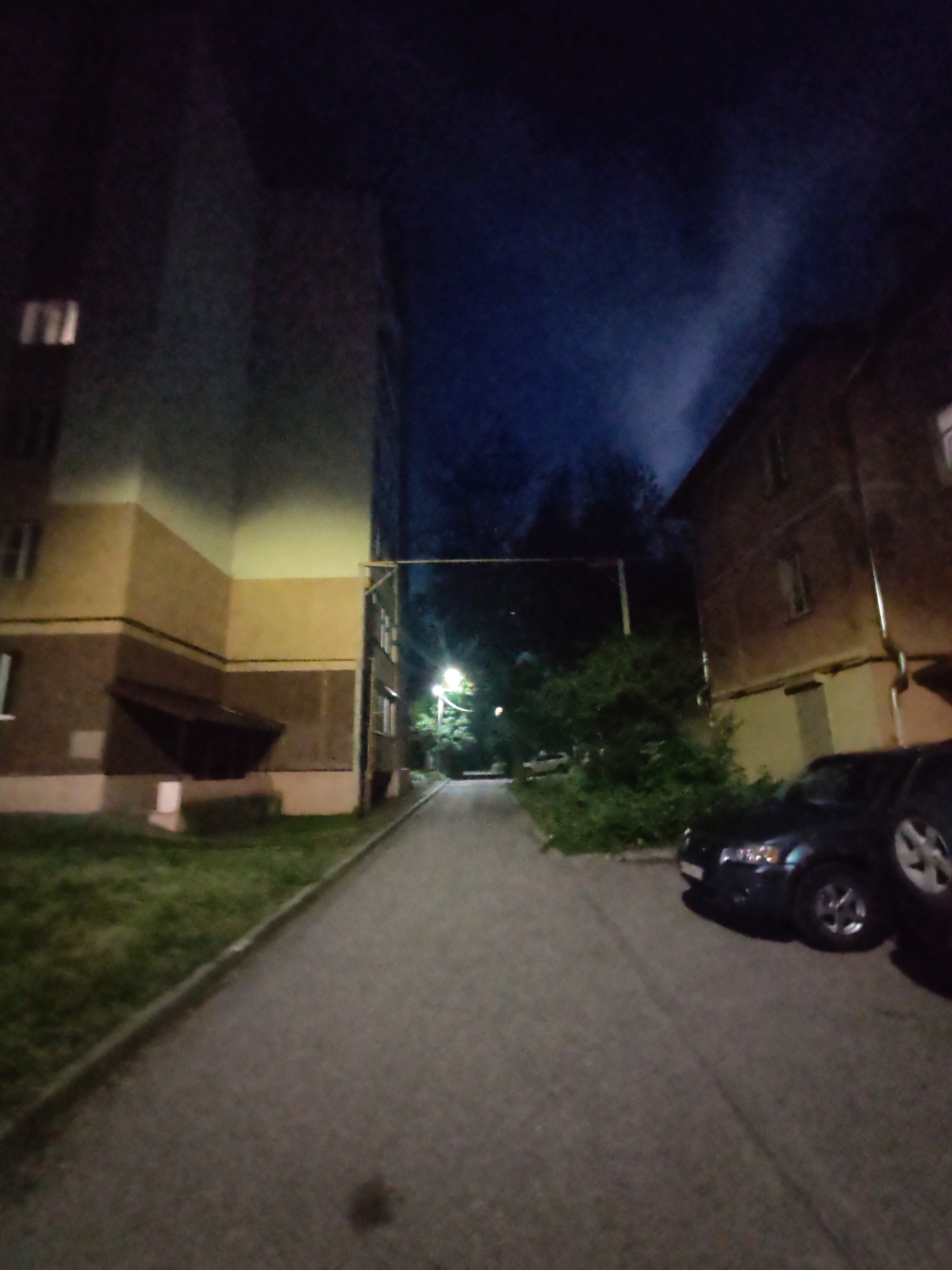A nighttime, low-light, medium-shot photograph depicts a view down a paved walkway between two apartment buildings. The building on the left is multi-story with a beige and brown facade, featuring multiple windows and an overhang. The building on the right is also multi-story, with a brown facade and visible gutters. Two dark-colored cars are parked on the right side of the walkway, one in the foreground and another partially visible behind it. The walkway is lined with grass on the left. A lamppost illuminates the scene with a warm light, casting shadows. The sky above is dark blue with faint wisps of light-colored smoke or clouds. The scene appears to be a residential area in the city of Vladimir, Russia, possibly in a courtyard or alley between buildings. No people are visible in the frame.