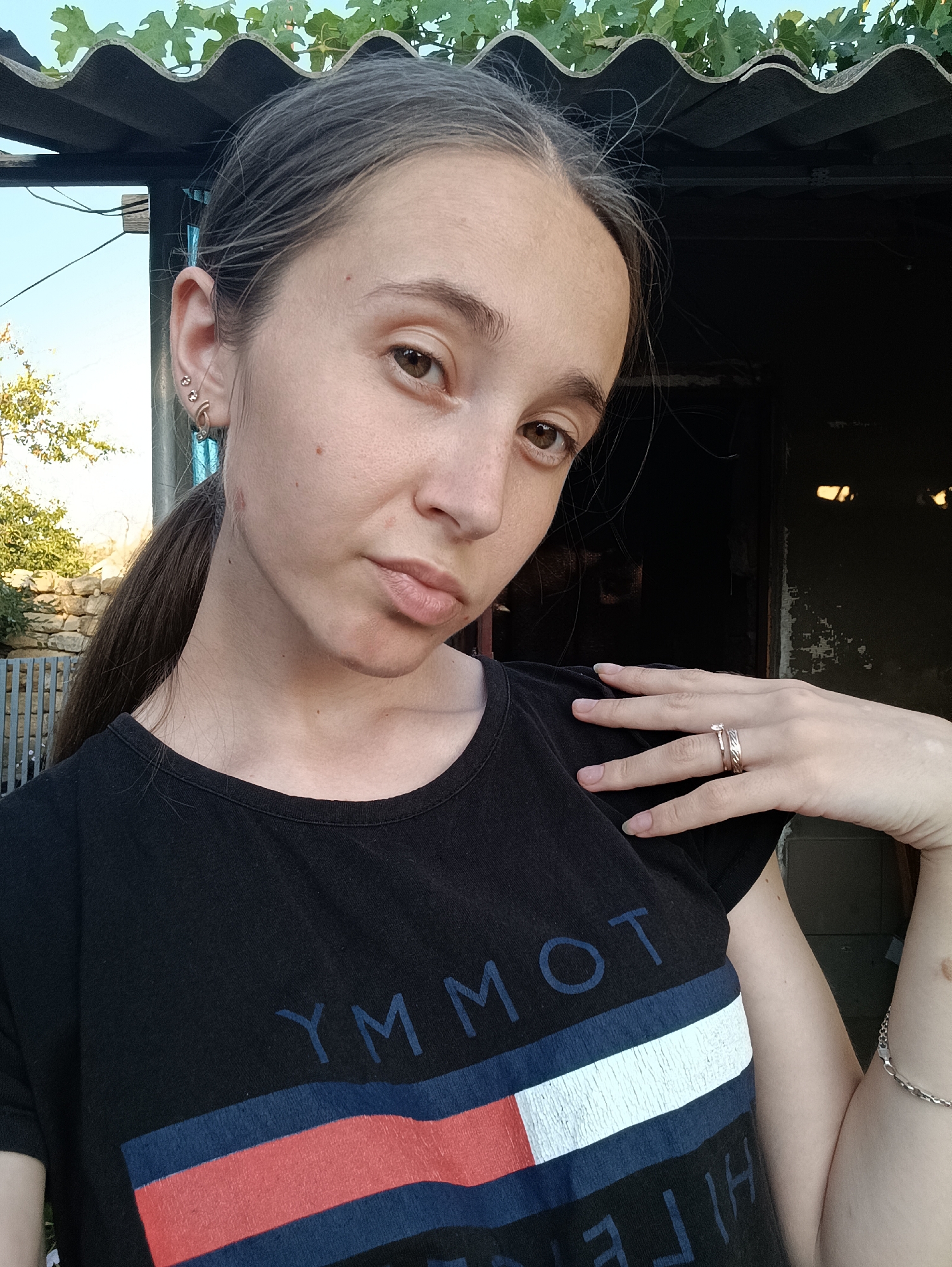 A young woman with brown hair pulled into a ponytail takes a selfie outdoors. She is wearing a black t-shirt with a Tommy Hilfiger logo. Her right hand is resting on her chest, displaying two rings on her finger and a bracelet on her wrist. She has two earrings in her left ear. The background shows a corrugated roof and a rustic building. The woman appears to be in a rural setting, possibly Vypasne, Ukraine.