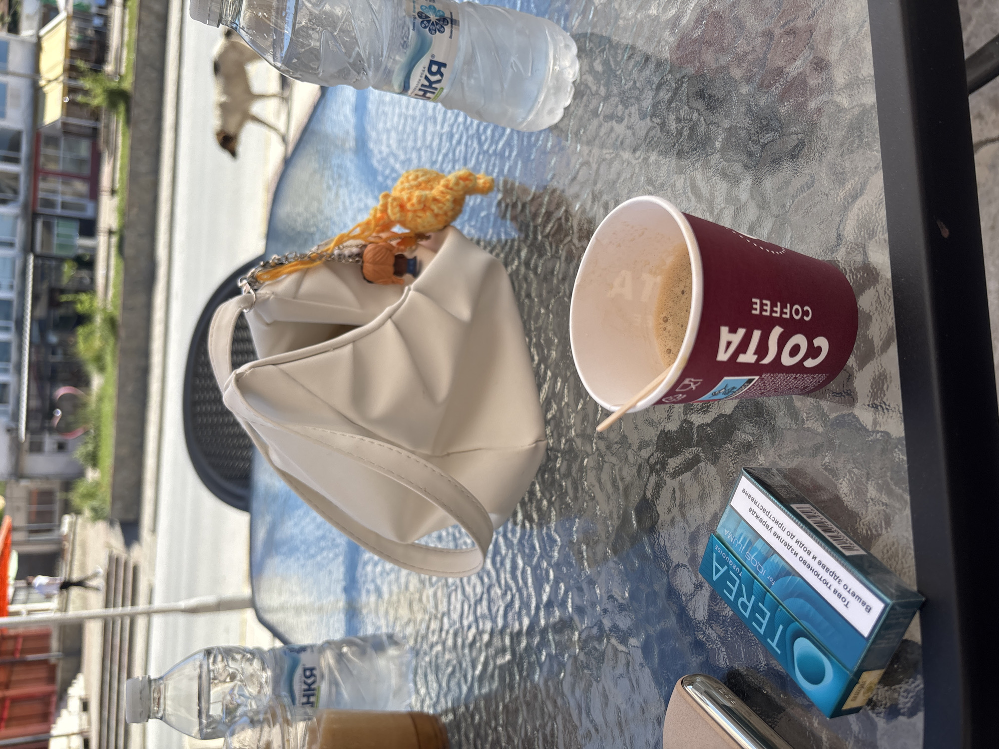 An outdoor table at a cafe in Garmen, Bulgaria, is set with a coffee cup, a pack of Terea Turquoise for IQOS Iluma cigarettes, a white purse with a keychain, and two bottles of water. A person is walking in the blurred background, and a dog is walking on the street. The table has a glass top with a textured pattern, and it is positioned on a patio overlooking a street with buildings and trees. The lighting suggests it is daytime.