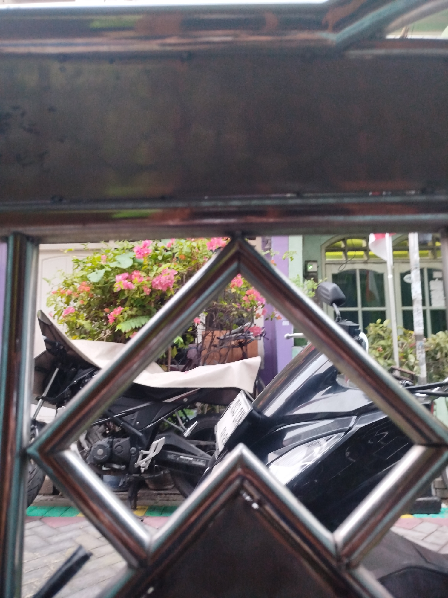 A motorcycle is parked in front of a building with pink bougainvillea in the foreground. The motorcycle is black with a white seat cover. The building has white doors and windows. The scene is framed by a metal grate. This is likely a residential area in Surabaya, Indonesia, given the architecture and vegetation.