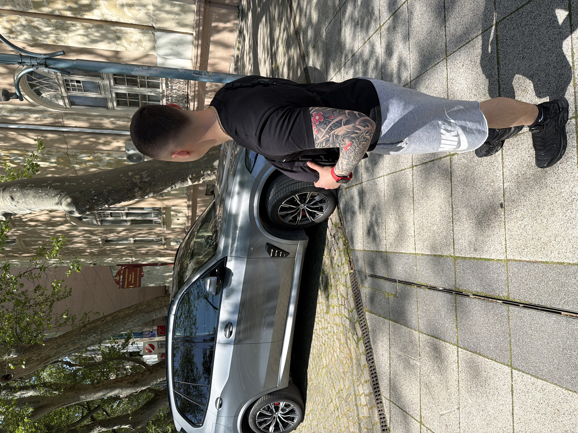 The image displays a full-body view of a man standing on a paved sidewalk next to a parked silver SUV, illuminated by bright daylight. The scene is set on a street in Plovdiv, Bulgaria, identifiable by a visible street sign.
The man, seen from behind and slightly to his left, has short dark hair. He wears a black short-sleeved t-shirt, grey athletic shorts with "NIKE" printed in white on the left leg, and black Nike Air Max Plus TN sneakers. A gold chain is visible around his neck, and a red wristband or smartwatch adorns his left wrist. His left arm is covered by a colorful tattoo sleeve featuring red cherry blossoms and a detailed dragon design.
Beside him, a silver SUV, likely a BMW X series (such as an X5 or X3 M Sport, indicated by wheel design and side vents), is parked. The vehicle has dark tinted windows. The ground beneath is composed of light grey paving tiles and darker cobblestones, with a long drain running parallel to the curb. Distinct shadows from trees and the man himself are cast on the ground, suggesting a sunny day.
In the background, an older building with multiple windows, including an arched one, stands under the dappled shade of a large tree. A street light structure is also visible. A red and white street sign, in Bulgarian, explicitly states "Паркирането разрешено" (Parking allowed) and specifies "Понеделник - Петък 08:00 - 18:00" (Monday - Friday 08:00 - 18:00). The man appears to be casually standing or waiting.