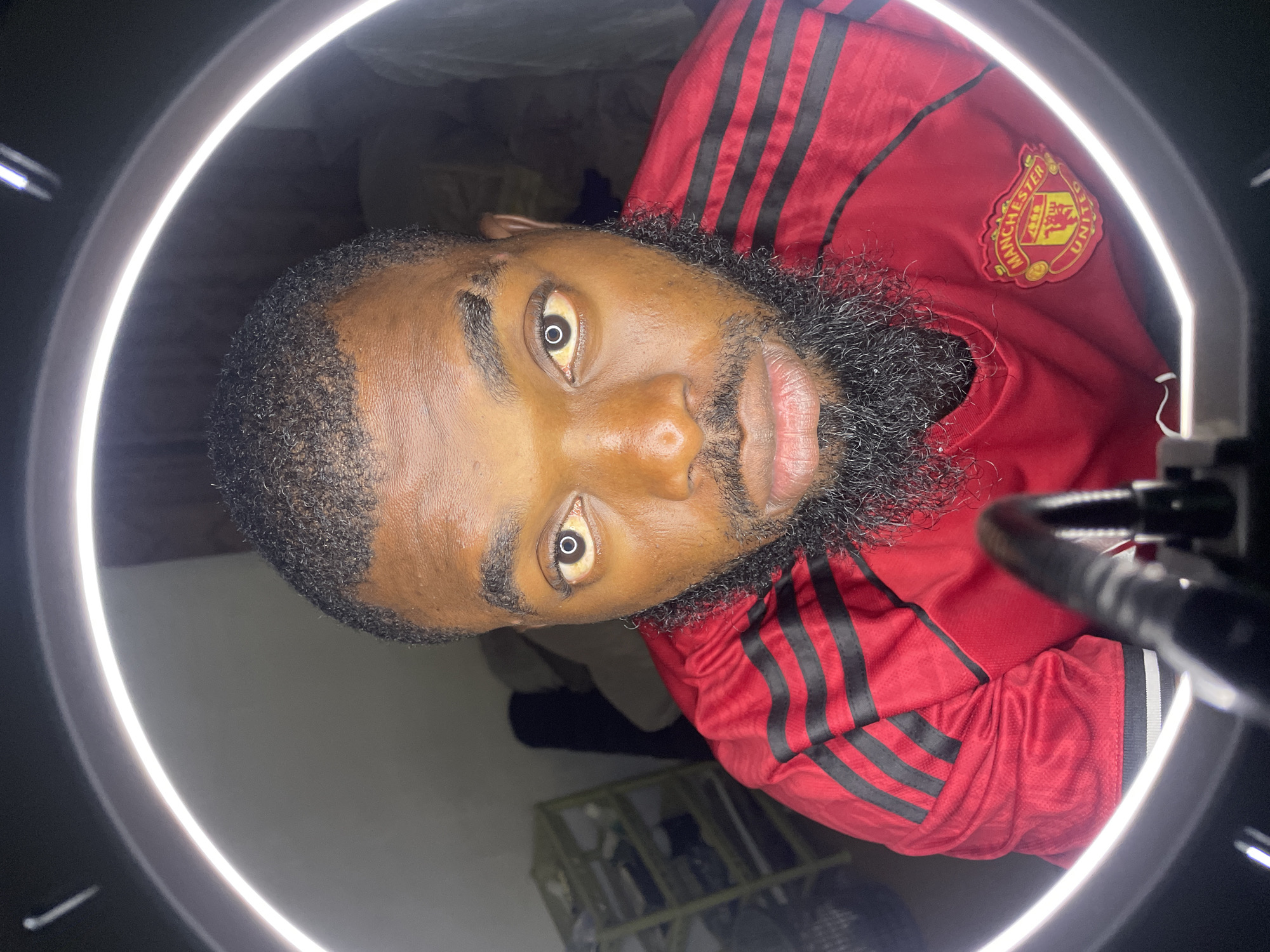 A man with dark skin, a beard, and short hair is looking directly at the camera. He is wearing a red Manchester United jersey with black stripes on the sleeves and a badge on the chest. The man is framed by a ring light, creating a circular highlight in his eyes. The background is dimly lit, with a shelving unit visible on the left and a blurry couch or bed on the right. The setting appears to be indoors, possibly a bedroom or living space. The image was taken in Jos, Nigeria.
