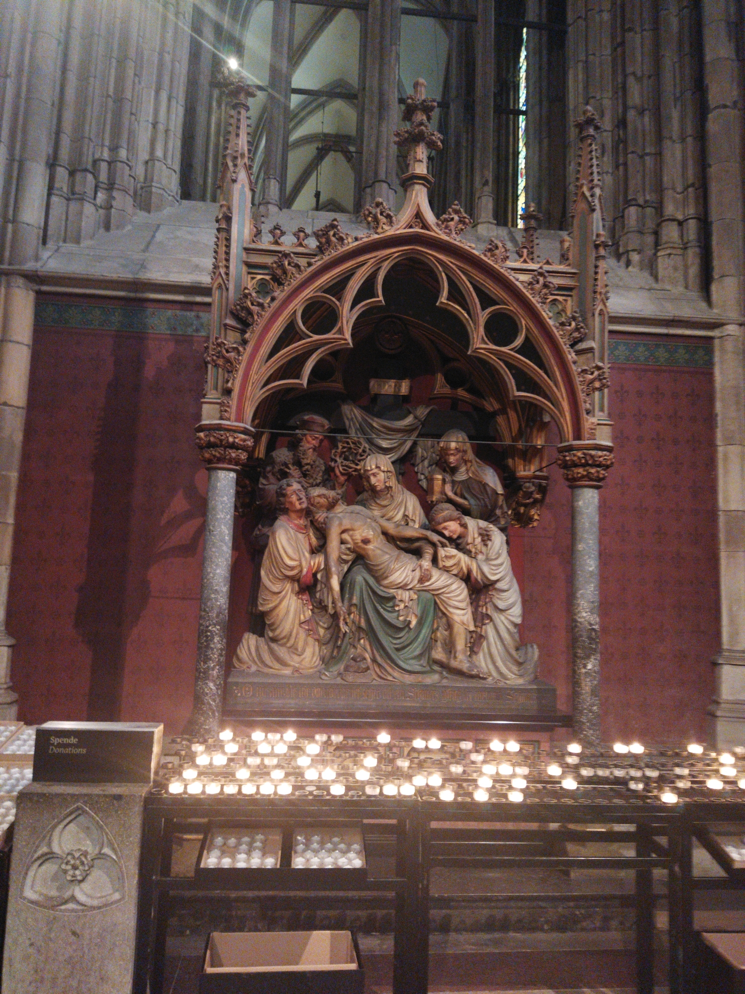 The image depicts a religious sculpture within a church. The sculpture, a pietà, shows the Virgin Mary cradling the body of Jesus Christ. Surrounding them are other figures, likely mourners. The sculpture is set within an ornate, Gothic-style niche made of wood and stone. In the foreground, there is a rack filled with numerous lit votive candles, some in glass holders and others directly on the rack. To the left of the candles is a stone pedestal with a decorative carving and a dark box labeled "Spende Donations". The background shows architectural elements of the church, including tall stone columns, arched windows, and a stained-glass window. The overall scene suggests a place of worship, likely a Catholic church, with an atmosphere of reverence and solemnity. The specific location within Monheim am Rhein, Germany, is not explicitly identifiable from the image itself, but it is a scene commonly found in historical churches.