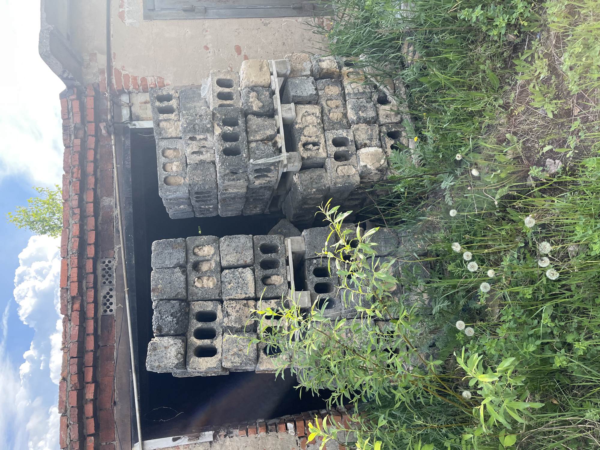 The image shows two stacks of grey concrete blocks with three rectangular holes each, resting inside what appears to be a partially collapsed brick structure. The blocks are stacked on top of wooden supports, visible between the layers. The brick structure is weathered and has missing bricks. To the right of the blocks is overgrown vegetation, including tall grasses and dandelions. To the left is a brick wall with deteriorated plaster. The scene is somewhat overgrown, suggesting a neglected or abandoned area. The location in the city of Chaikovsky, Russia, is not evident in the photo.