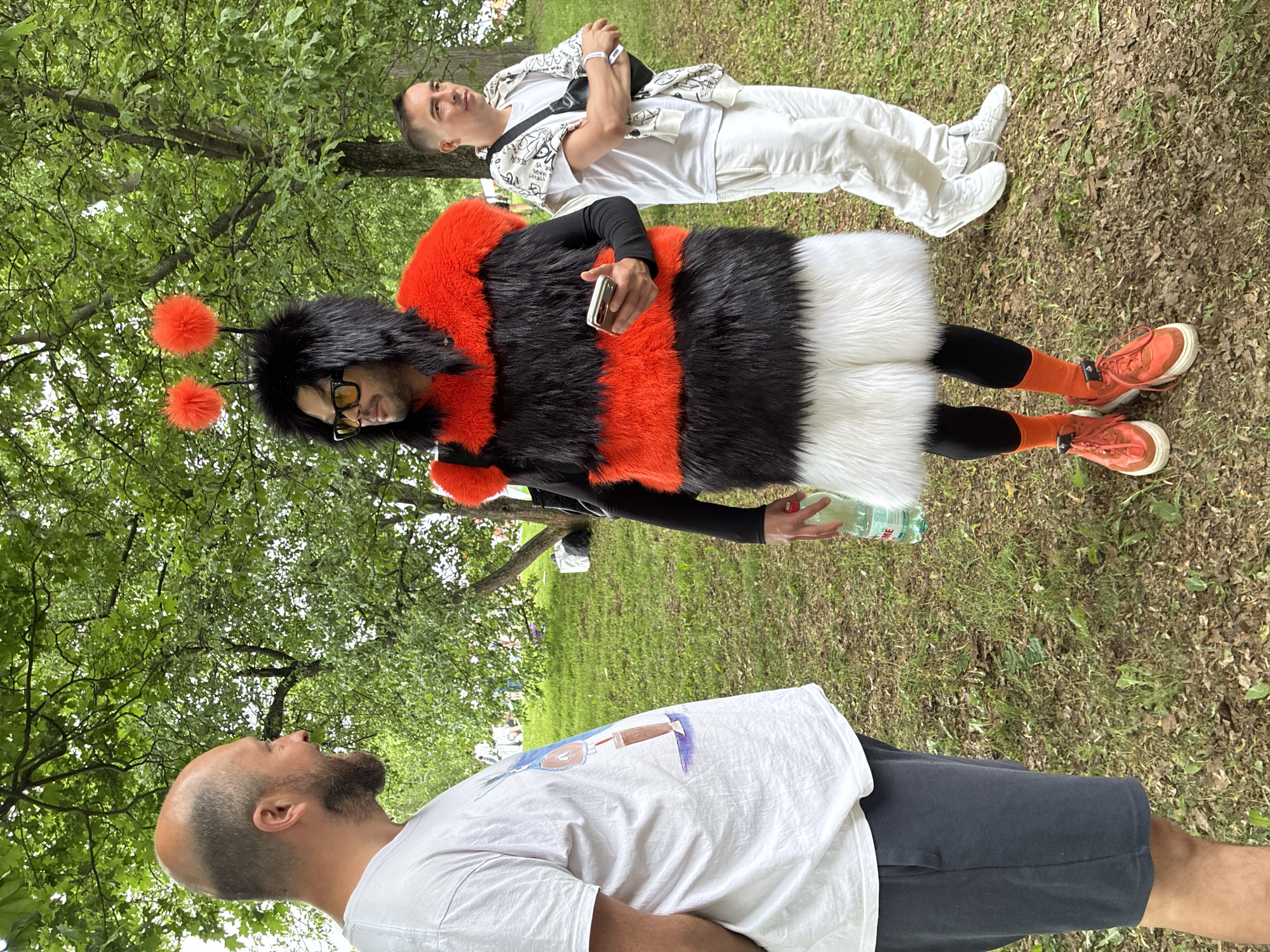 The image shows three men standing in a grassy park area. The man in the center is wearing a bumblebee costume consisting of horizontal bands of orange, black and white faux fur, black leggings, orange socks, and orange sneakers. He also has a black wig with orange pom-poms and glasses. He is holding a smartphone in his left hand and a water bottle in his right. Another man is lying on the ground behind him, dressed in a white outfit with a patterned white jacket and a black bag across his body. He has a wristband on his right arm. The third man is in the foreground, wearing a white t-shirt and black shorts. He is bald and looking up toward the trees. Green trees provide a background to the scene. The exact location in Moscow, Russia cannot be determined without more information.