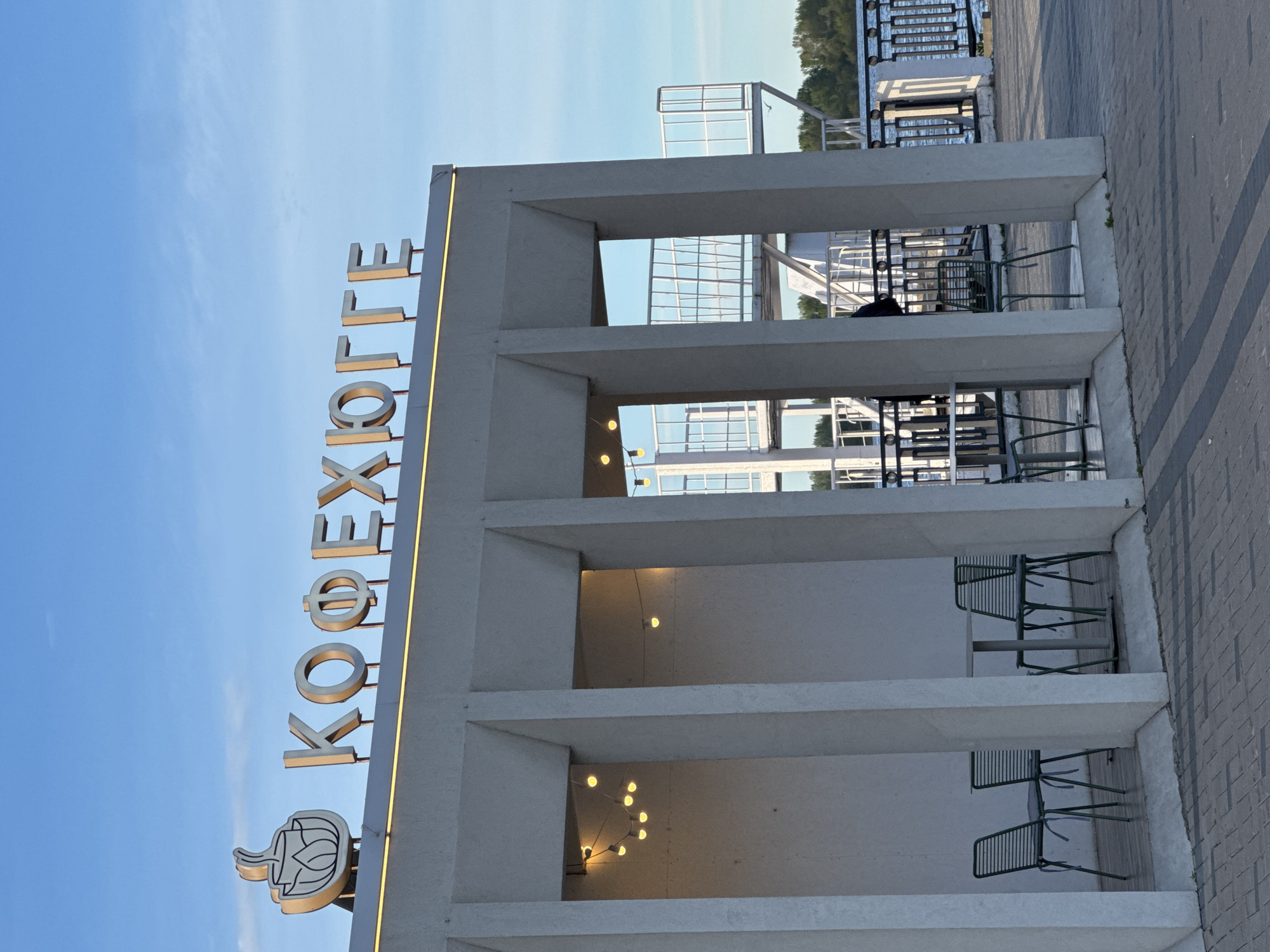 The image shows the Koexolle restaurant, located in Nizhny Novgorod, Russia. The vertical sign "KOEXOLLE" is prominent on the left side of the building's facade. The architecture consists of rectangular concrete sections, creating open, framed spaces. These spaces contain tables and chairs. The background includes a partial view of a river or body of water with a metal railing. The sky is visible above the building, showing a gradient from blue to a lighter color near the horizon.
