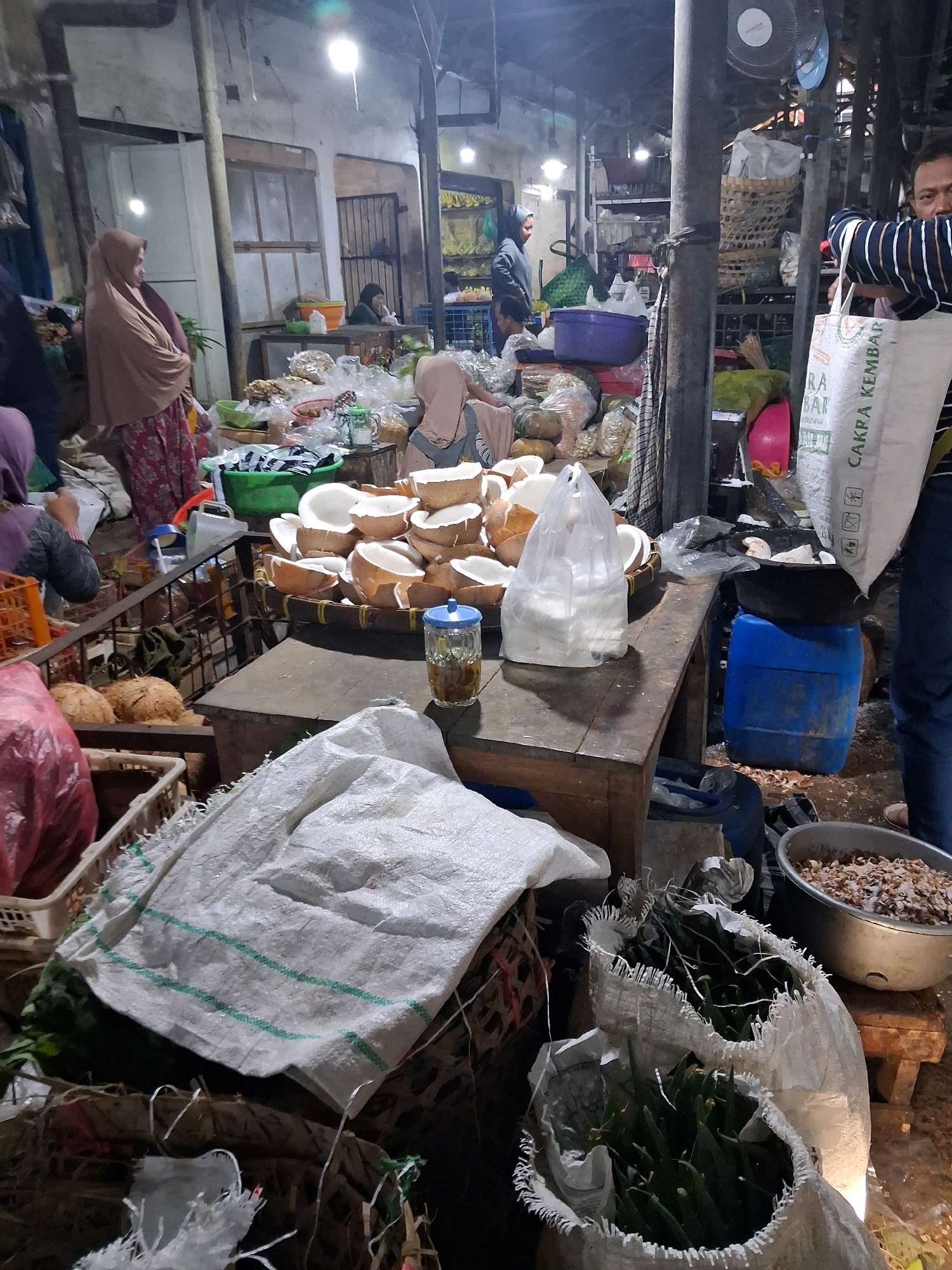 The image depicts a bustling market scene in Surakarta, Indonesia, likely a traditional market. In the foreground, a white woven bag with green stripes lies on top of other woven materials. To the right, a silver bowl contains dried food items, possibly seeds or nuts. Next to it, another white woven bag is filled with green, elongated vegetables, possibly chilies or some form of bean.
Further back, a wooden table is laden with various items. A clear glass jar with a blue lid holds a yellowish liquid, likely oil or a drink. Next to it, a transparent plastic bag contains white shredded material, possibly coconut. A woven basket holds a stack of halved coconuts.
In the middle ground, several individuals are visible. A woman in a light brown hijab and a patterned skirt stands facing away from the camera. Another person wearing a light brown hijab is seated behind the table, partially obscured. To the left, a person in a purple hijab is also partially visible. A man wearing a striped shirt is holding a white bag with text on it, facing the camera.
The environment appears to be an open-air market or a covered but rustic structure, with exposed beams and overhead lighting. Various other goods, including what appears to be produce and packaged items, are visible in the background, suggesting a diverse marketplace. A blue barrel is situated behind the table. The overall impression is one of a vibrant, active market with a variety of goods and people engaged in commerce.