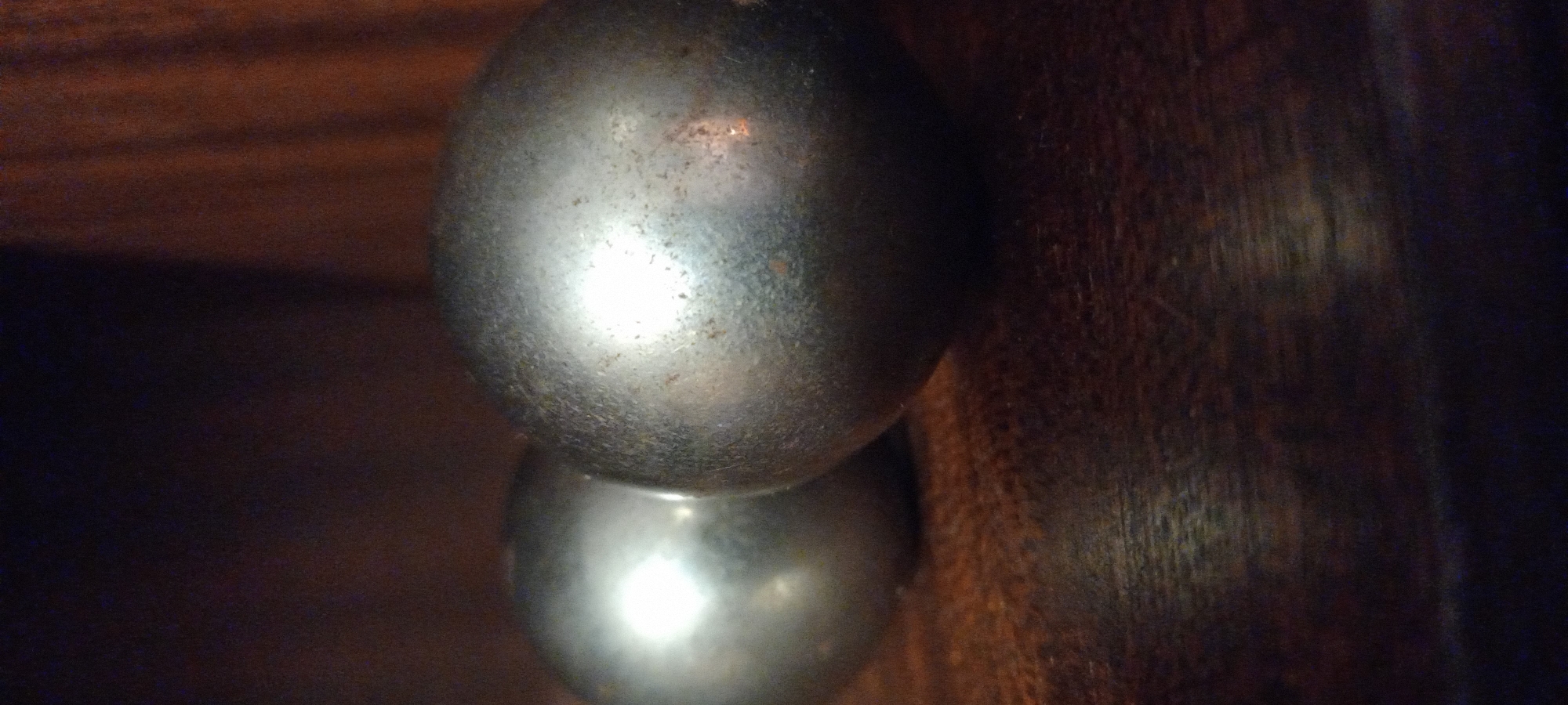 The image presents a close-up, vertically oriented shot of two spherical, metallic objects set against a dark, textured wooden background. The objects appear to be tarnished or aged silver-colored spheres, possibly doorknobs or decorative finials, reflecting a bright light source in their centers, creating strong highlights. The top sphere is almost entirely visible, showing a slightly uneven surface with specks of rust or dirt. The bottom sphere is partially visible, appearing directly below the first and also displaying a bright reflection.
The background on the left is blurred and dark, while the right side and top feature a rich, dark brown wooden surface with visible grain and texture, possibly a piece of furniture, a door, or a wall panel. The lighting is dim overall, emphasizing the metallic sheen and reflections of the spheres. There is no activity taking place, as it is a still life focused on these objects. No people are present, and no text is visible. The scene suggests an indoor setting, likely in low-light conditions, with a direct light source illuminating the metallic spheres. The overall aesthetic is dark and textural.