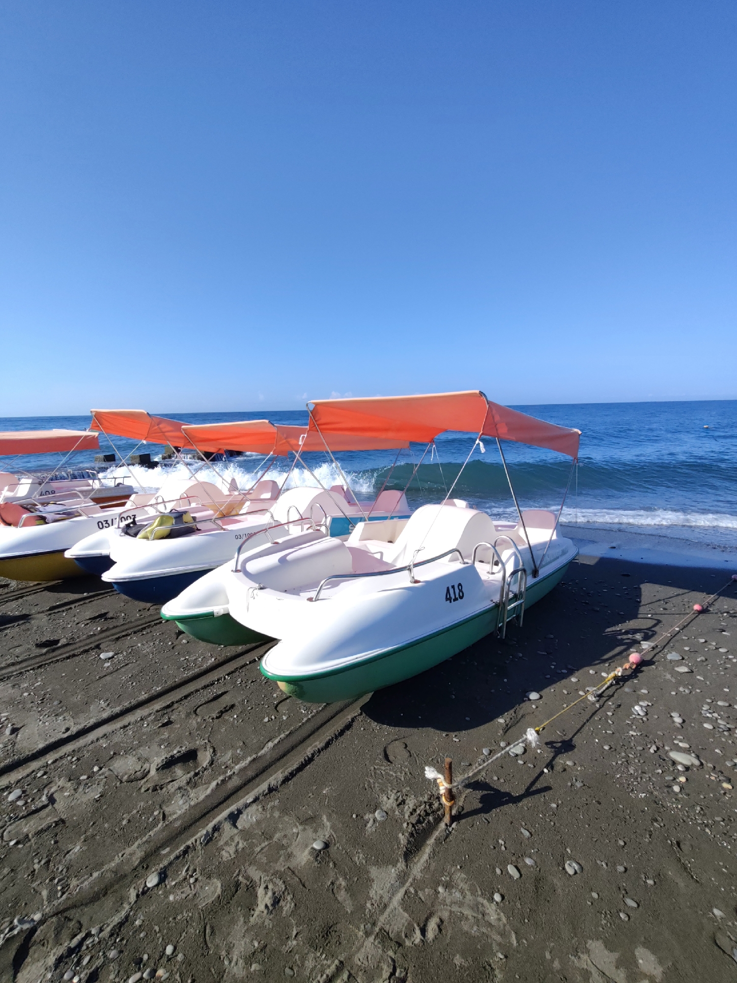 A row of white and green pedal boats with orange canopies are lined up on a dark, pebbly beach. The boats are positioned on tracks in the sand, with the closest boat on the right having the number "418" visible on its side. The sea, a deep blue, is calm with gentle waves breaking on the shore. The sky is clear and a vibrant blue. The scene appears to be a beach resort area, likely in Sochi, Russia, given the commonality of such recreational activities in that region. There are no people visible in the image. The overall impression is one of a peaceful, sunny day at the seaside.