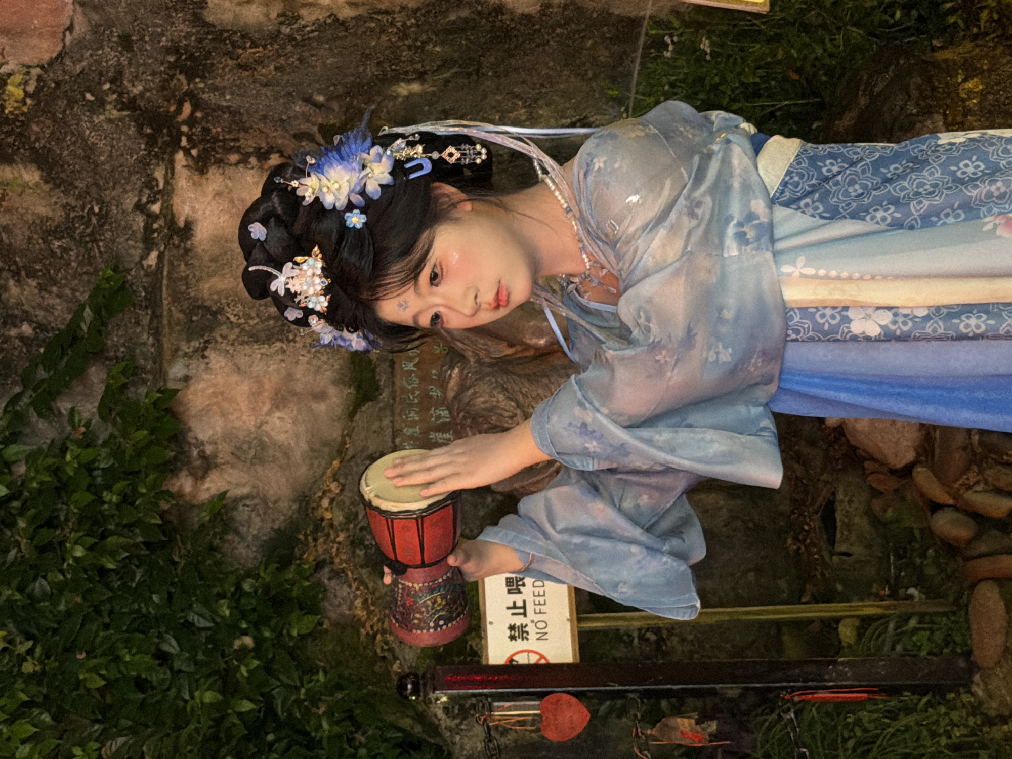 A young woman in traditional Chinese attire, including a blue floral patterned robe and an elaborate headdress adorned with blue flowers and pearls, plays a small red and tan drum. She is positioned outdoors against a textured stone wall partially covered in greenery. A sign with Chinese characters and "NO FEED" is visible behind her. The lighting suggests it is either evening or a shaded area. The woman's makeup includes rosy cheeks and a decorative mark on her forehead. The overall scene evokes a sense of traditional culture or performance. The location is specified as Yuzhong District, China.