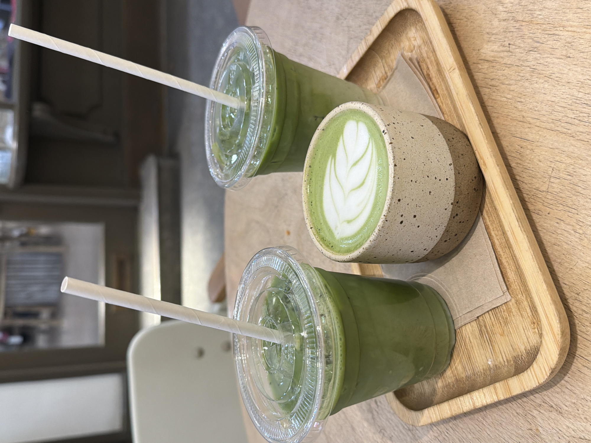 The image shows a close-up of a wooden tray with three drinks. Two of the drinks are in clear plastic cups with lids and white straws, containing a green liquid, likely iced matcha lattes. The third drink is in a ceramic cup with a latte art design on top, also a shade of green, likely a hot matcha latte. The tray is sitting on a wooden table. The background is blurred, but suggests an interior space, possibly a cafe. Given that the image is cropped and does not contain a significant portion of the location, it is impossible to ascertain if the location is in the city of London, United Kingdom.
