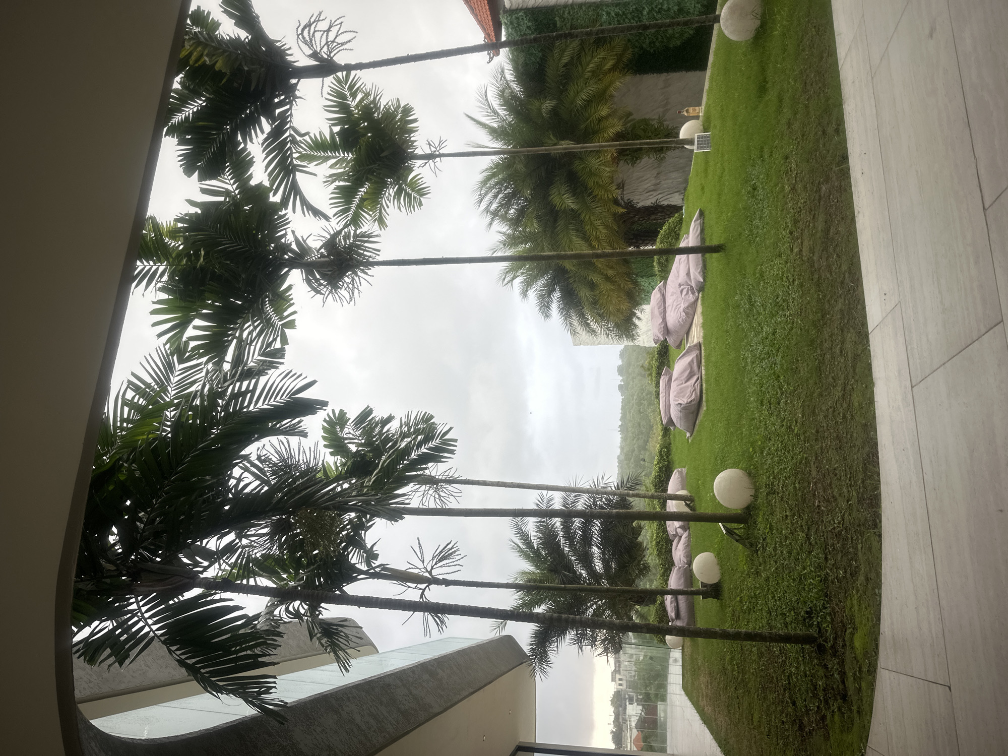 The image depicts an outdoor garden space in Kuta Selatan, Indonesia, as seen through a window or opening. Several palm trees line the edge of a grassy area where numerous pink beanbag chairs are placed. Spherical light fixtures are interspersed along the lawn. In the distance, buildings can be observed. The sky is overcast, suggesting a cloudy day. The angle of the shot indicates a view from an elevated position.