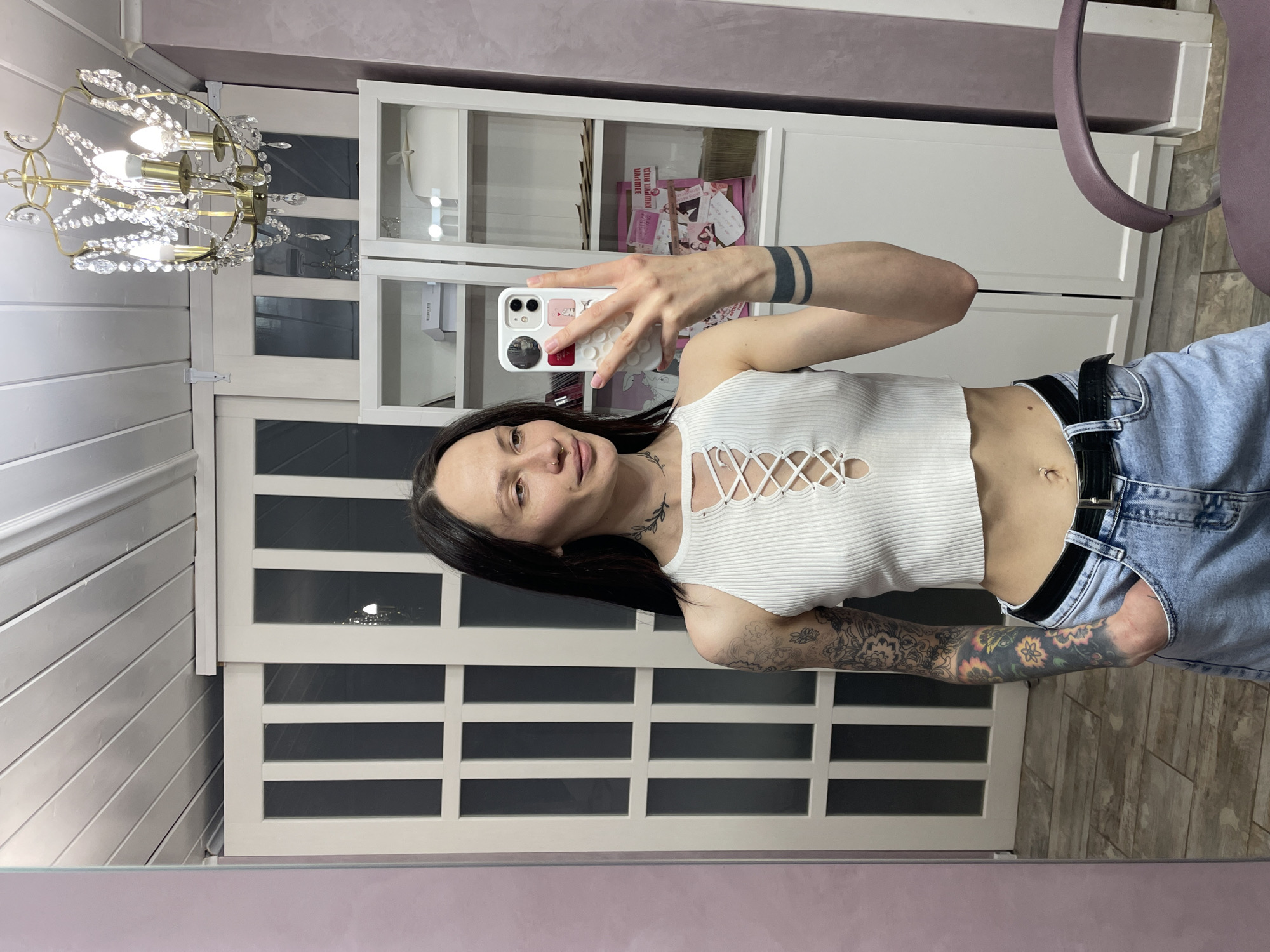 A woman is taking a selfie in a room. She has dark hair and tattoos on her arm and neck. She is wearing a white crop top and light blue jeans with a black belt. She is holding a white smartphone with a patterned case. Behind her is a white cabinet with glass doors and a white wall with decorative panels. A chandelier is visible in the upper left corner. It is not possible to determine the location as Izhevsk, Russia with the given information.
