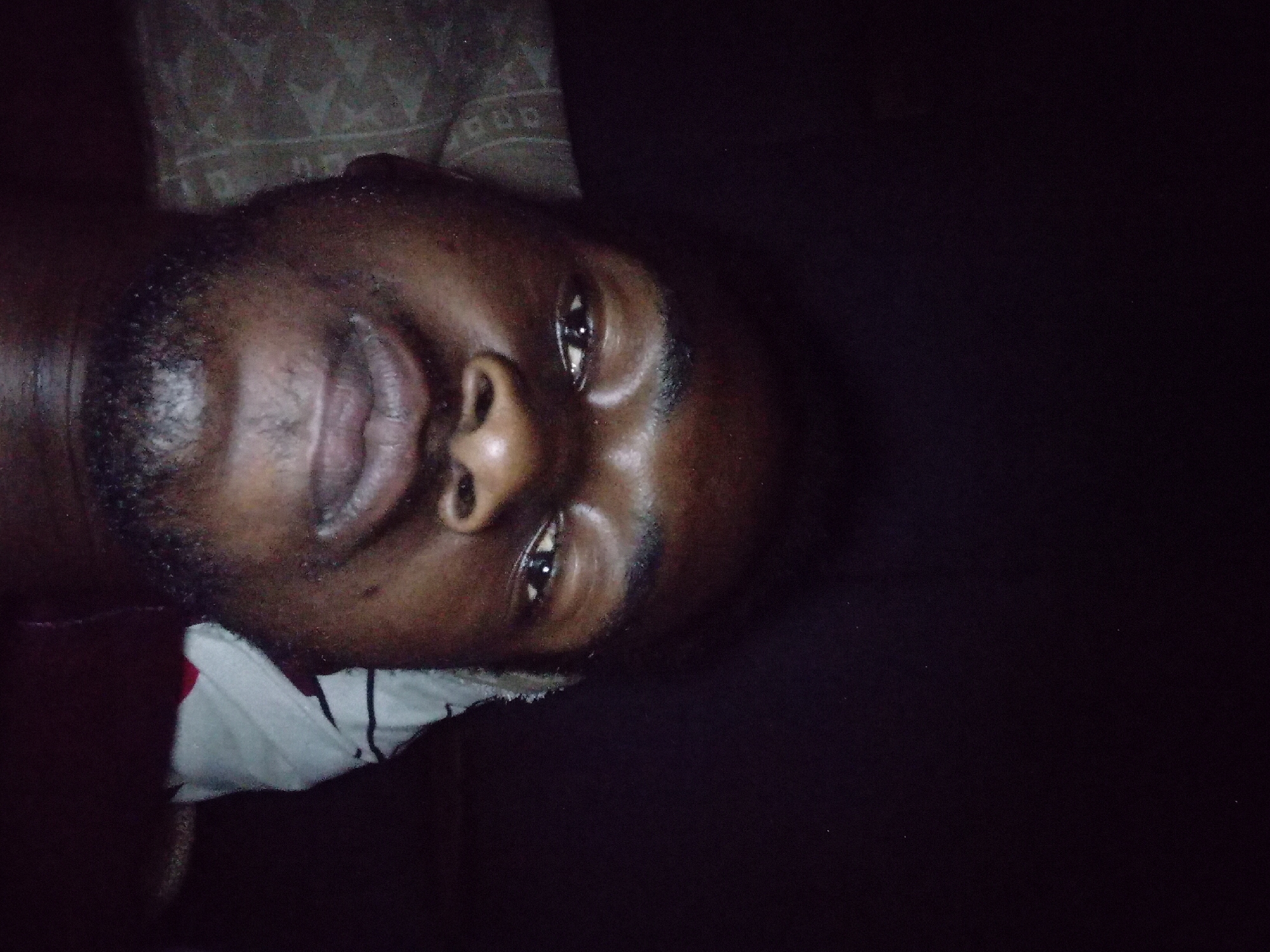 A close-up, low-light portrait captures a dark-skinned individual, likely male, appearing to be lying down and looking directly into the camera. The face, featuring dark eyes, a broad nose, and full lips, is centrally positioned in the lower half of the frame. Short, dark facial hair forms a beard and mustache around the mouth and chin. Reflections are visible on the forehead, nose, and lips, indicating a direct light source. To the left of the individual's head, a section of a light-colored, patterned pillow or bedding is visible. On the right, near the neck, a white garment with dark horizontal stripes is partially visible, layered over a darker, possibly red or maroon fabric. The background and the upper portion of the image are predominantly dark, with subtle image noise, suggesting a dimly lit indoor environment.