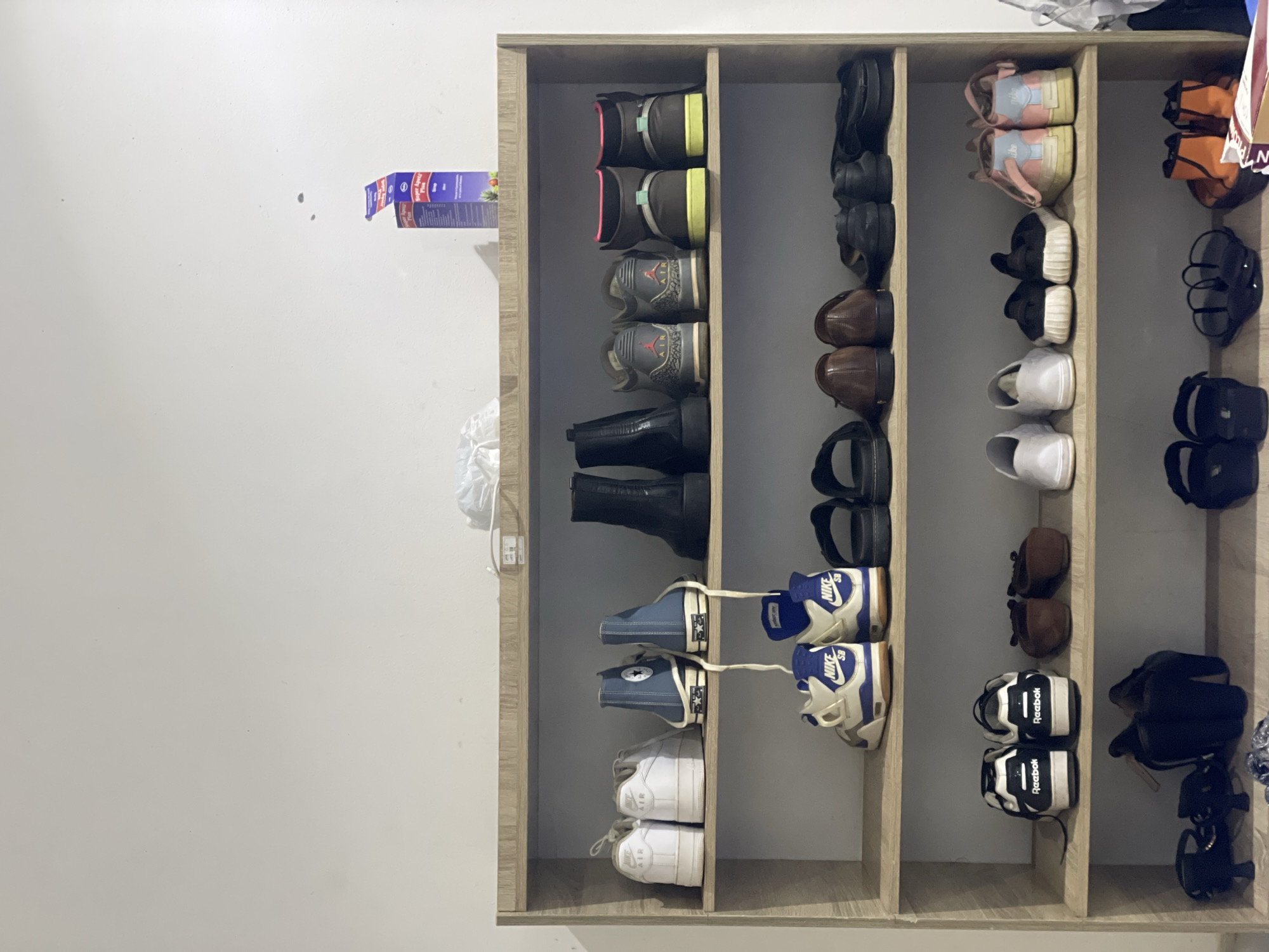 The image shows a light-colored wooden shoe rack filled with various pairs of shoes. The rack has five shelves. The top shelf has a box of medicine and some crumpled white material. The second shelf from the top contains two pairs of white sneakers, two pairs of blue high-top sneakers, and two black boots. The third shelf has two pairs of white and blue sneakers, two pairs of brown loafers, and two pairs of black slip-on shoes. The fourth shelf has two pairs of black and white Reebok sneakers, two pairs of brown flat shoes, two pairs of black shoes, and two pairs of pink and blue sneakers. The bottom shelf has two pairs of black sandals, two pairs of black flip-flops, and two pairs of orange and black boots. The background is a plain white wall. The scene is indoors, likely a home, and there are no people visible. The presence of the shoe rack and the variety of footwear suggest a personal collection of shoes. No specific details about the city of Abuja, Nigeria, are present in the image itself.