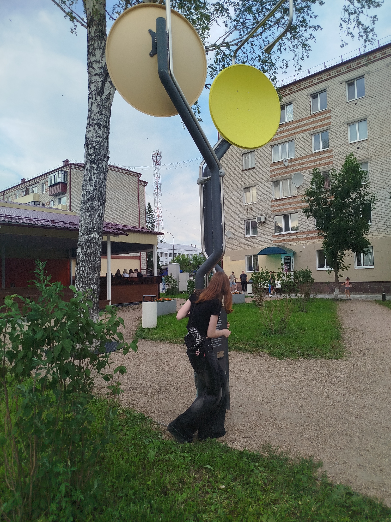 In Zavodoukovsk, Russia, a young woman with long, reddish-brown hair is captured in a full-body shot, facing away from the camera and slightly to the left. She is wearing a black t-shirt and dark, wide-leg pants, with a black and white checkered backpack on her back. Her left hand is reaching out and touching a black metal pole that is part of a sculptural installation. This installation features two large, circular discs; one is pale yellow and the other is bright yellow. The woman is standing on a gravel path. To her left, a tall birch tree with distinctive white and black bark stands prominently, with its branches extending towards the sky. In the background, a multi-story brick apartment building with light-colored brick and red striped accents dominates the scene. In front of the apartment building, there is a covered outdoor seating area with people seated, and further to the right, a green tree. A cellular tower is visible in the distance. The environment appears to be an outdoor public space, possibly a courtyard or park, with patches of green grass and shrubs. The sky is overcast.