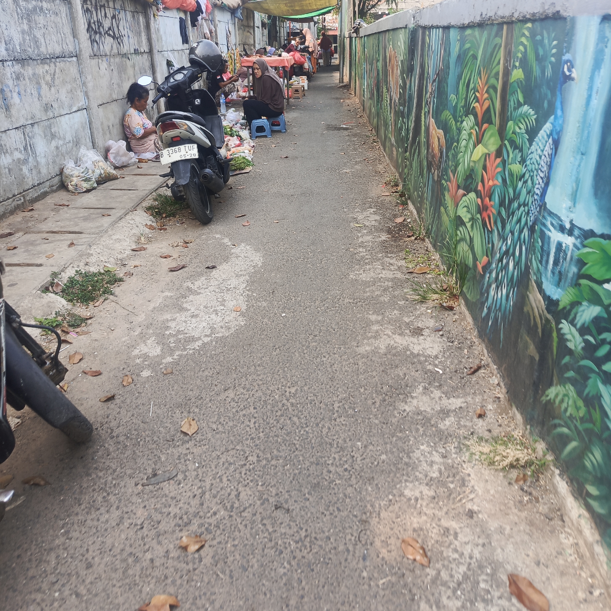 This image depicts a street scene in South Jakarta, Indonesia, possibly near a market or residential area. A narrow asphalt path runs through the scene, bordered on the left by a concrete wall covered in graffiti and on the right by a vibrantly painted mural of a jungle scene featuring animals like deer and a peacock, and plants like gingers and ferns.
On the left side of the path, near the graffiti-covered wall, a woman sits on the ground next to a motorcycle. She is wearing a light-colored patterned top and appears to be looking down. Several plastic bags filled with produce or goods are piled next to her. A white and black motorcycle with a helmet on the handlebars is parked on the path.
Further down the path, a second woman wearing a brown hijab and dark clothing sits on a blue plastic stool next to a stall. She is surrounded by various items, possibly for sale, including produce and other goods. A small white cat is visible near the front wheel of the motorcycle. The path is littered with fallen leaves, and patches of grass and weeds grow along the edges. Clothes hang from a line above the stalls in the background, suggesting a living or working space adjacent to the market area. The overall impression is of a bustling, informal urban environment.
