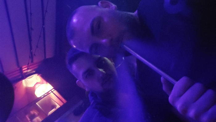 The image depicts two males in a dimly lit, purple-illuminated setting, likely an indoor or semi-outdoor lounge in Plovdiv, Bulgaria. The foreground male, with a shaved head and facial stubble, holds a dark hookah hose to his mouth and exhales a visible plume of white vapor. He wears a dark shirt. Positioned behind and to his left, a second male, also with a very short hairstyle, looks towards the camera. He wears a dark top. In the upper left background, a warm, glowing light fixture with a light-colored shade is visible. An overhead structure, possibly a ceiling or canopy, features horizontal lines and what appear to be string lights.