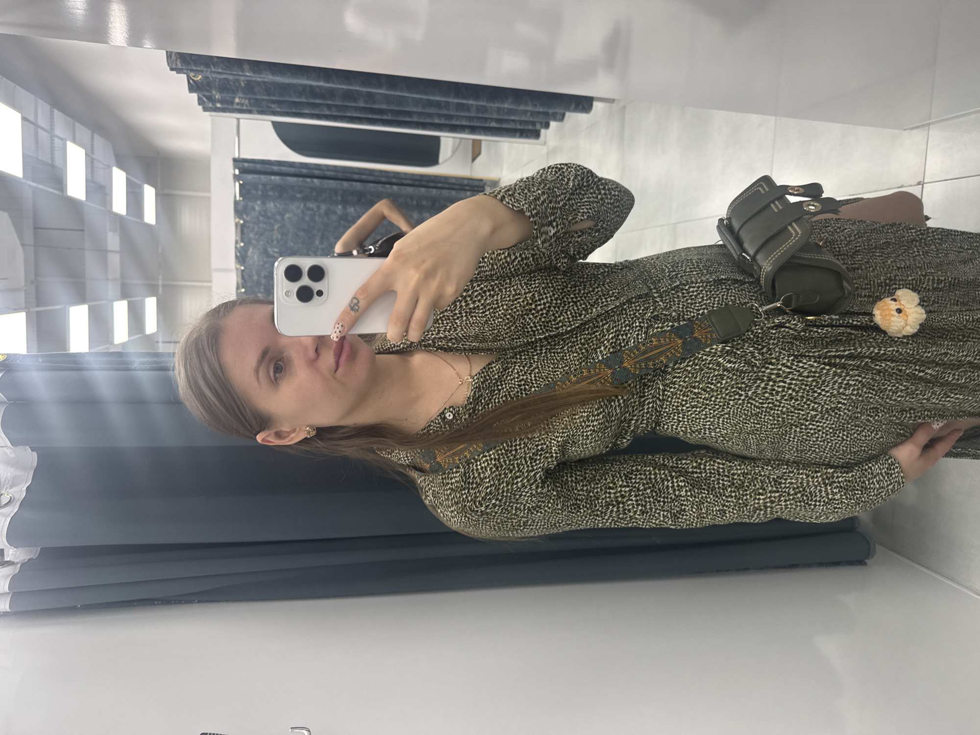 A woman is taking a selfie in what appears to be a fitting room. She is holding a white smartphone with multiple cameras. She is wearing a patterned green dress with a dark green handbag slung across her body. The environment includes clothing racks with curtains and the bright lights typical of retail spaces. The location is a dressing room. There is no information about a city or Batumi, Georgia.