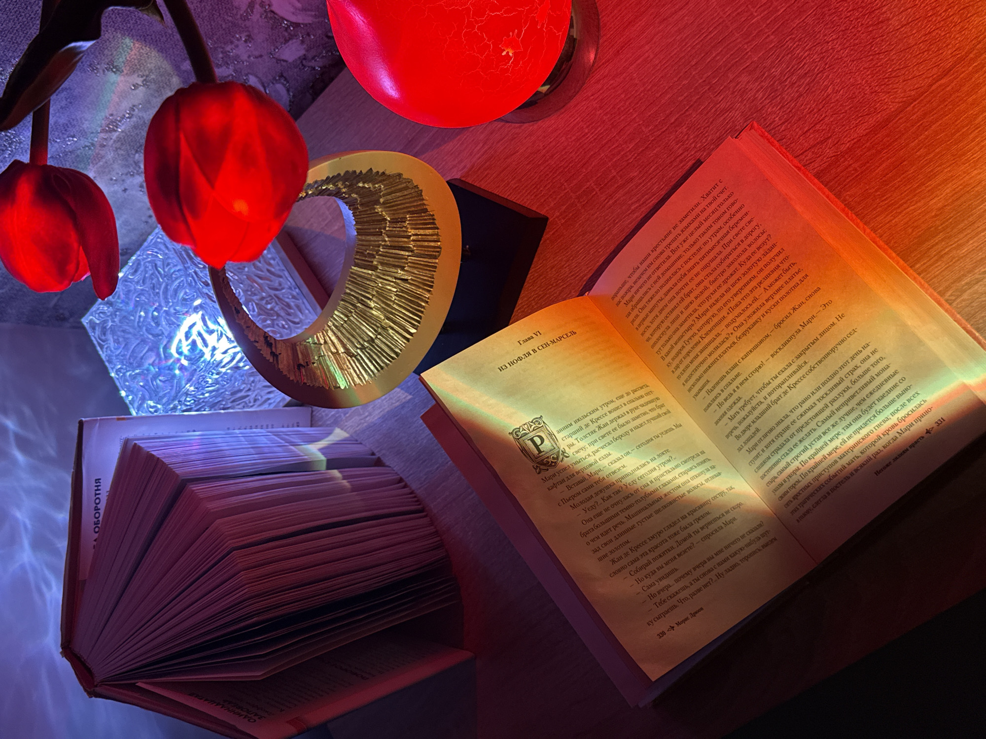 This indoor still life image features an open book surrounded by warm and cool lighting elements. In the foreground, a large, opened book with numerous pages lies flat, displaying text in Russian. The right page shows "Глава VI" (Chapter VI) and the chapter title "ИЗ НОФЛЯ В СЕН-МАРСЕЛЬ" (FROM NOHFL TO SAINT-MARCEL). At the bottom of the left page, "330 Морис Дрюов" (330 Maurice Druon) is visible, indicating a page number and author. On the right page, "Негоже лилиям прасть 331" (It is not fitting for lilies to spin 331) appears at the bottom. Another book, possibly closed or partially opened, is visible on the left, with text "А ОБОРОТНЯ" (A WEREWOLF) and partial words "ОДИННАА" (ELEVEN) and "ЗАПОВЕД" (COMMANDMENT / COVENANT) on its edge/spine.
The scene is dimly lit by artificial sources, creating a cozy and atmospheric mood. To the left, two red, glowing tulip-shaped lights hang downwards, casting a warm red hue. To their right, a golden, ribbed, crescent-shaped decorative object reflects light. A vibrant red, cracked-texture candle, also glowing, is positioned in the upper right. Below the hanging tulips, a reflective surface with a wavy or patterned texture glows with cool blue and purple light, contrasting with the warm tones. The overall lighting suggests an evening or nighttime setting, perfect for reading.