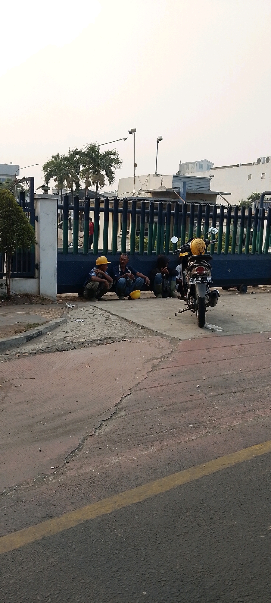 The image shows a street scene in front of a large gate, possibly an industrial compound, in the city of Bekasi, Indonesia. Three men are squatting on the ground near the gate, appearing to be taking a break. A motorcycle is parked to the right of the men, with a yellow helmet resting on the back seat. Palm trees are visible behind the gate, and buildings can be seen in the background. The ground is paved with asphalt and concrete, showing some signs of wear and tear. The sky is overcast, suggesting a cloudy day. The overall atmosphere of the image is one of everyday life and work in an urban environment.