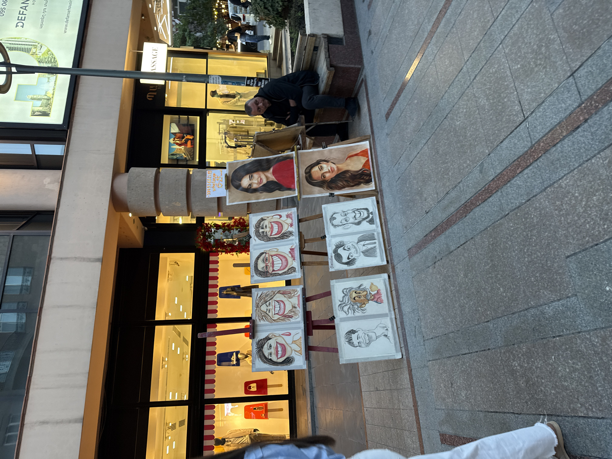 Here is a description of the image:
The image shows a street scene in Yerevan, Armenia, near a "Massage" store. An artist sits on a bench, leaning over an easel displaying his art. The artist has black clothing. A selection of portrait and caricature artwork is displayed on multiple small easels on the sidewalk in front of a storefront with illuminated window displays of purses and handbags. A sign in Armenian is visible with a price list. The sidewalk is paved with gray stone tiles, interspersed with bronze trim.