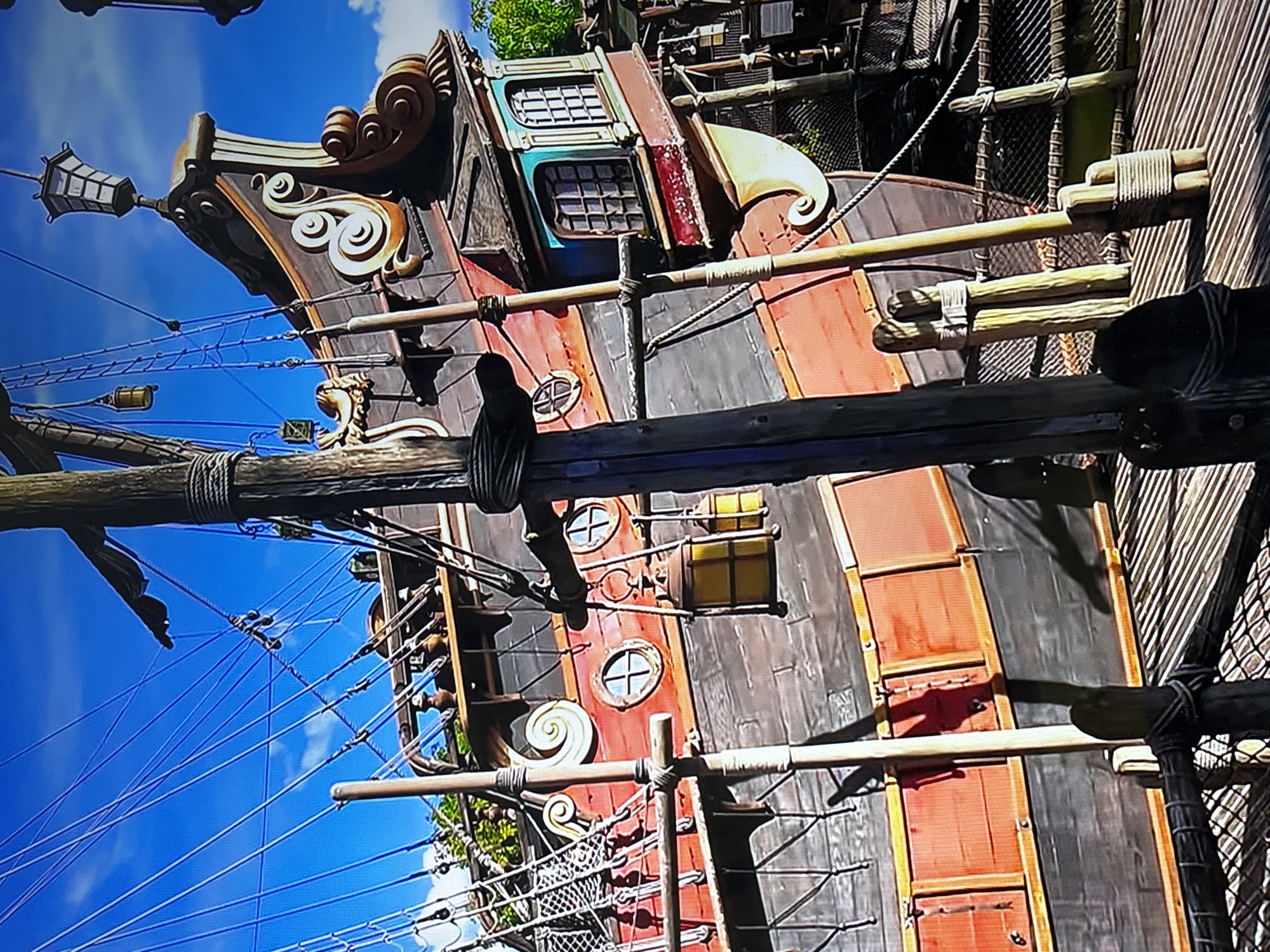 A partial view of a pirate ship with a dark red and black hull under a partly cloudy blue sky. The ship has decorative white scrolls, porthole windows with crosses, and a lantern at its stern. A tall wooden mast and rigging are visible against the sky. Wooden structures and rope netting partially obscure the right side of the ship. The location is Pirates of the Caribbean, Disneyland.