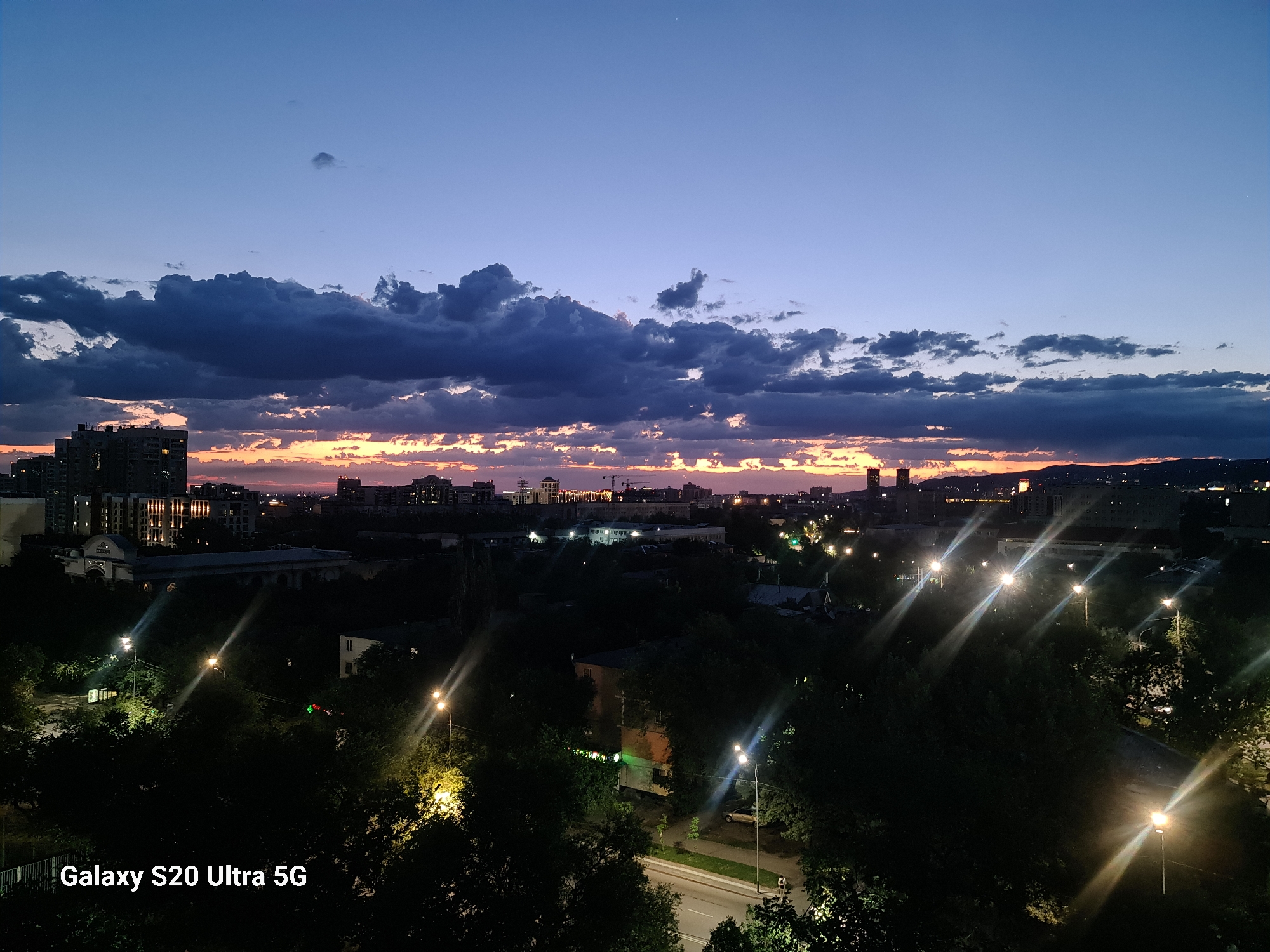 The image captures a twilight scene over the city of Almaty, Kazakhstan. The sky is a gradient of deep blue at the top, transitioning to lighter hues of purple and orange near the horizon where the sun has recently set. Dramatic, dark clouds are scattered across the upper portion of the sky, with breaks revealing the warm glow of the sunset.
Below the sky, the cityscape is visible. Tall buildings, some appearing to be apartment blocks, are silhouetted against the colorful sky. The mid-ground is densely populated with trees, their dark forms punctuated by the artificial lights of streetlamps and building interiors. These streetlights cast long, starburst-like flares across the image, a common effect in nighttime photography with bright light sources. A road is visible, also lined with trees and illuminated by streetlights. In the foreground, more trees are prominent, with the text "Galaxy S20 Ultra 5G" subtly watermarked in the bottom left corner. The overall impression is of an urban landscape at dusk, with the natural light fading and artificial lights beginning to dominate the scene. No people are clearly discernible.