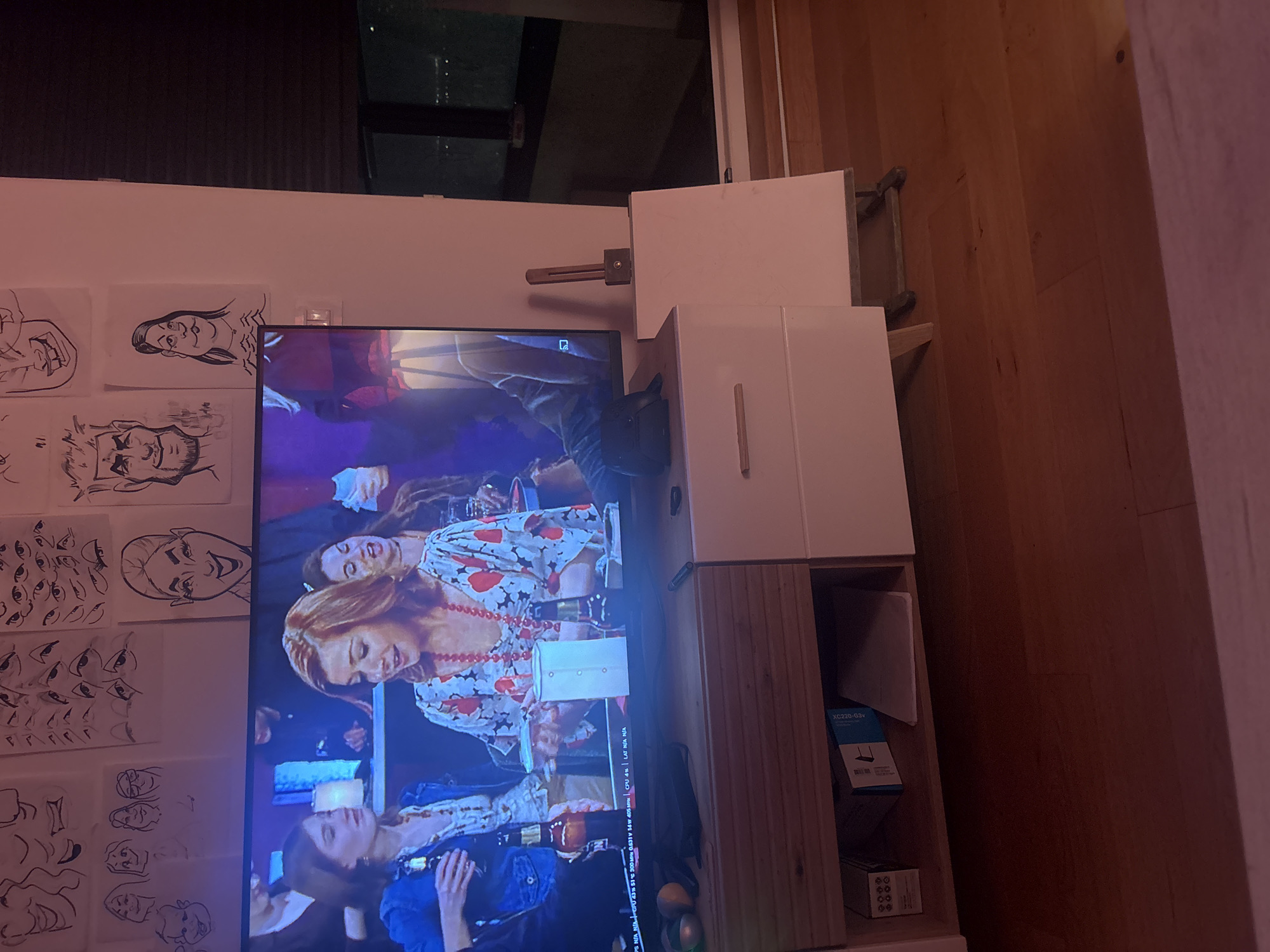 This is an indoor shot depicting a living space featuring a television set, artwork, and furniture. The television screen displays images of several women. Adjacent to the television is a wall decorated with hand-drawn sketches of faces and eyes. The furniture includes a white storage unit, where various items, including a box with the label "XC220-G3v" can be seen. The room is illuminated by overhead lighting, and the overall color palette is predominantly white and light brown. The presence of a window suggests a possible location in a city, however, the city of Belgrade, Serbia, cannot be confirmed from this image.