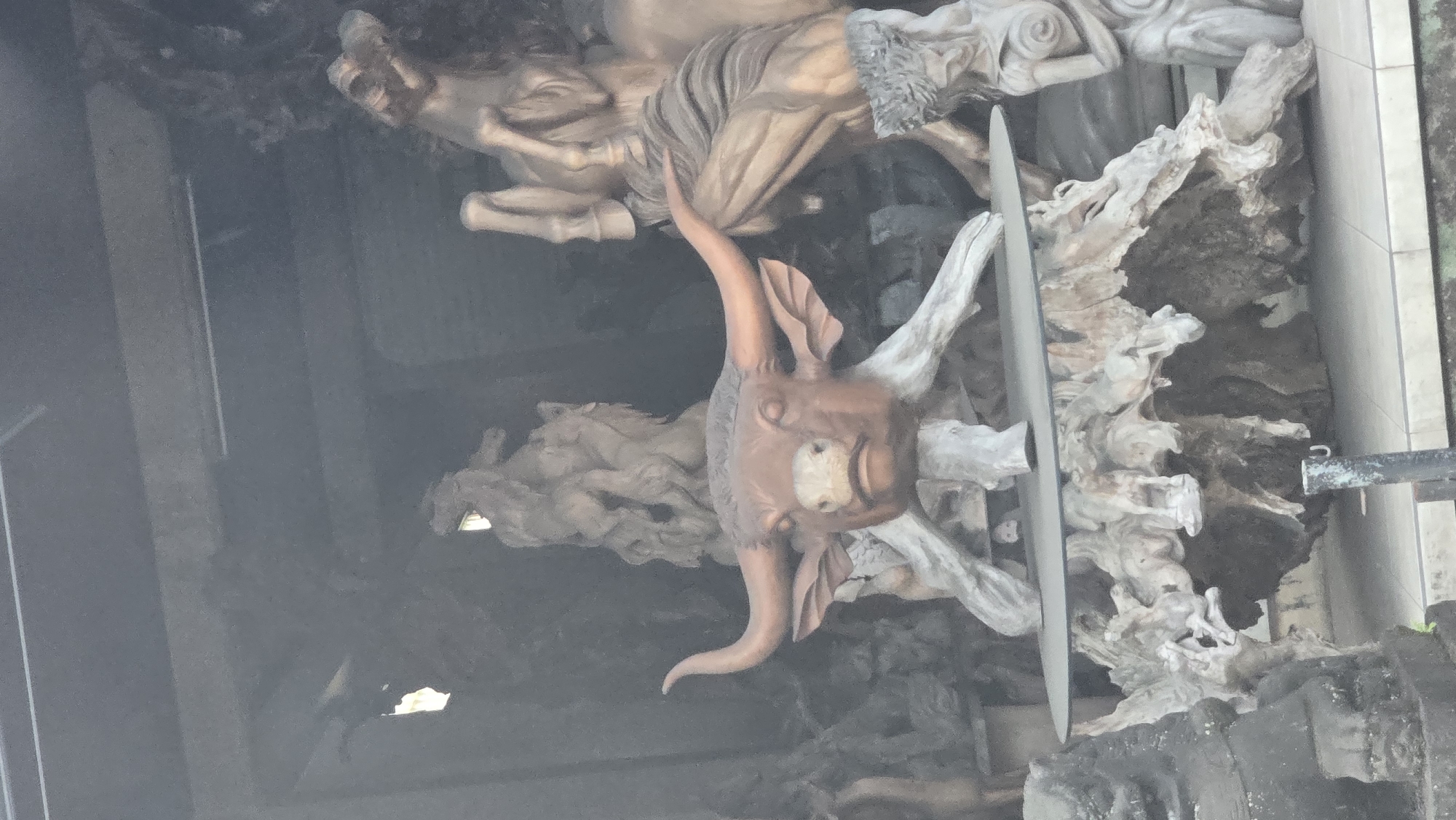The image features a close-up view of a sculpted installation in Ubud District, Indonesia. A central, brown-toned sculpture depicts a horned creature, possibly a demon or mythical being, with prominent curved horns and a simplified facial structure. Above and behind this central figure, there is a larger sculpture featuring human figures and horses, appearing in a lighter, more weathered tone. The sculptures are set against a backdrop of what seems to be simulated tree roots or rocky formations. A narrow, metallic, blade-like element slices through the central part of the composition, obscuring part of the horned creature and the tree roots. The overall setting appears to be an outdoor, public space, judging by the partially visible wall tiles and the presence of a metallic structure in the background.