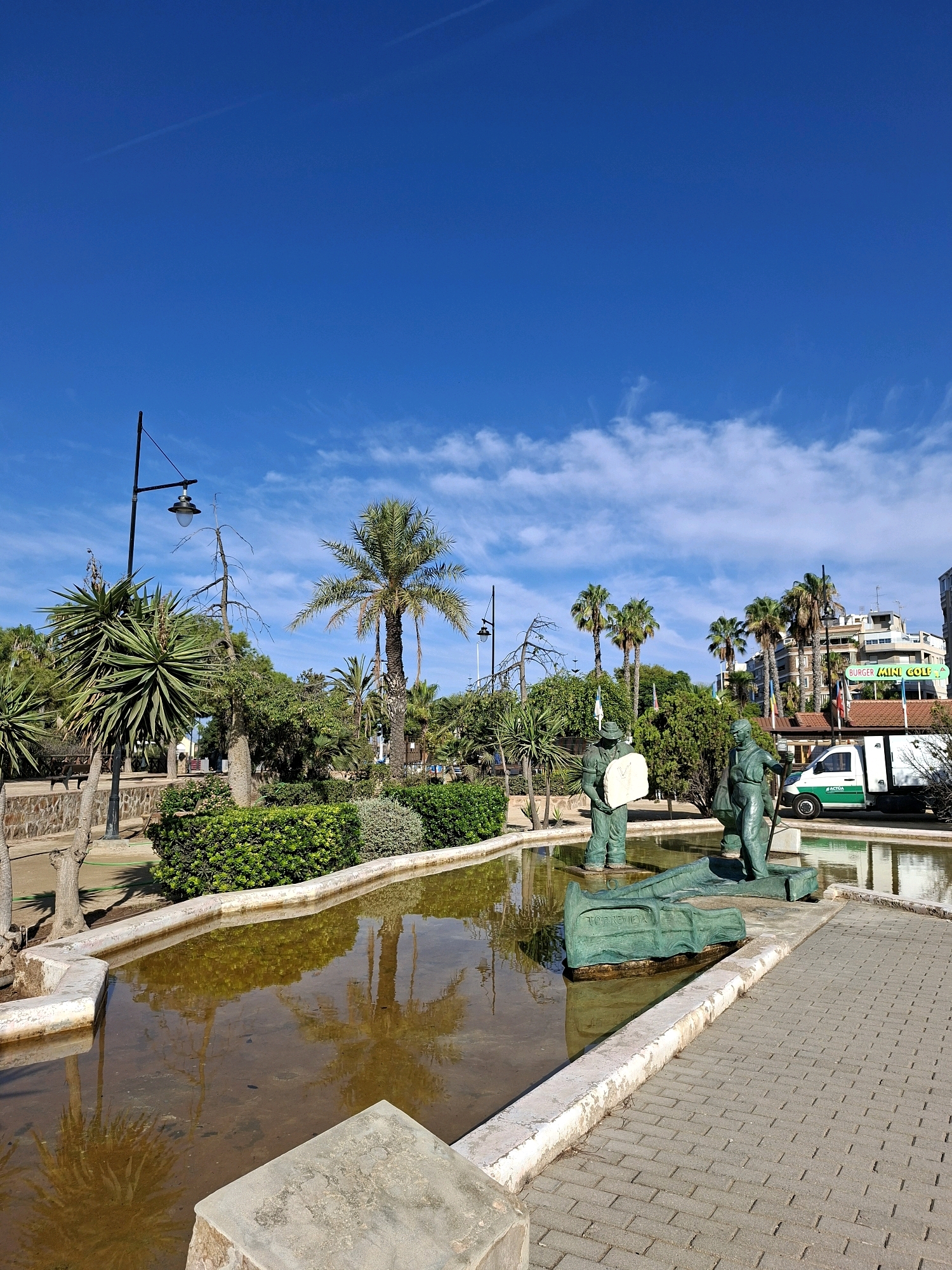 The image depicts a public park in Torrevieja, Spain, featuring a shallow water fountain with bronze statues. The statues appear to represent people engaged in some form of labor or activity, with one statue holding a large, rectangular object. Another statue stands next to a long, green object that resembles a boat or a salvaged item, with the word "TORREVIEJA" inscribed on it.
The fountain is surrounded by a paved walkway made of interlocking bricks. Lush green bushes and several palm trees are visible around the fountain, creating a tropical ambiance. In the background, buildings and a white truck with green lettering are present. The sky above is a clear, bright blue with a few wispy clouds. The overall scene suggests a sunny day in a well-maintained urban park.