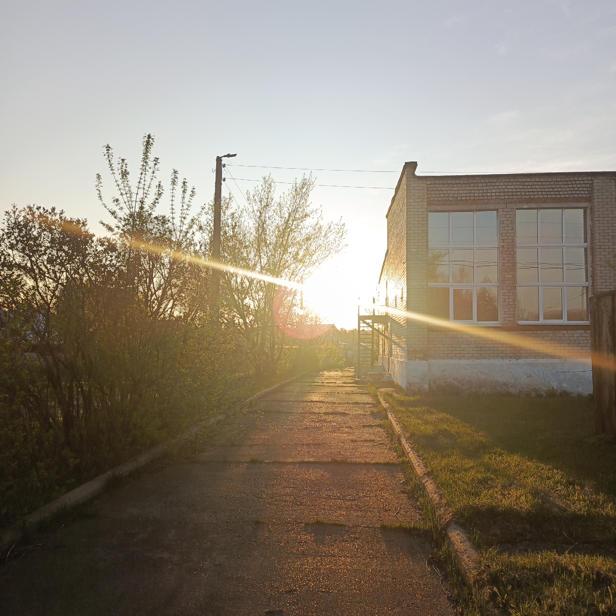 The image shows a street scene in Chaikovsky, Russia at sunset. A concrete path stretches into the distance, illuminated by the setting sun. To the left of the path is a dense bush, with a wooden utility pole behind it. To the right, a brick building with two large windows is visible. The windows reflect the sky and some of the surrounding vegetation. The sky is a gradient of pale blue to orange near the horizon. There are no people visible in the scene.
