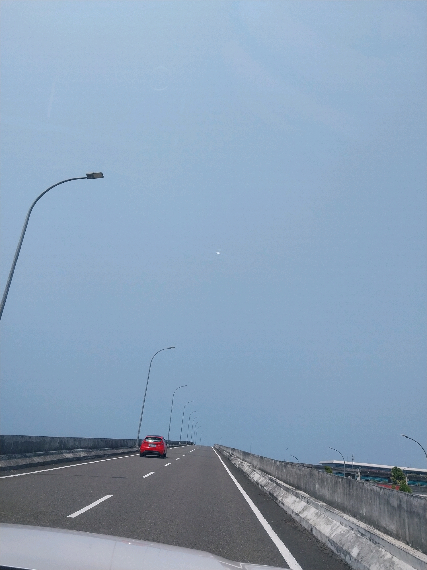 A red car is driving on a highway with a clear blue sky. Streetlights line the highway, and there are concrete barriers on either side. In the background, there is a building that appears to be a terminal or station. The scene is likely captured from a moving vehicle. The location is identified as the city of Glagah, Indonesia.