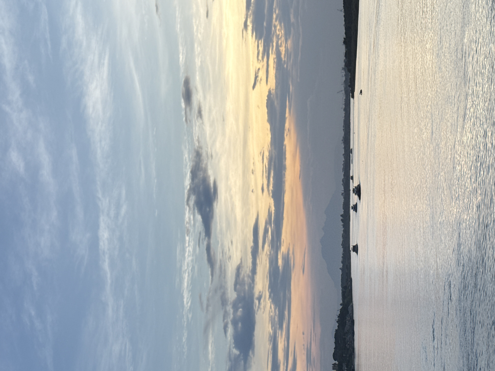 The image shows a body of water, likely a lake or river, reflecting the sky with boats in the distance. The sky is filled with clouds and a mix of blue, gray, and golden hues, suggesting sunrise or sunset. The shoreline on the left is lined with vegetation and possibly buildings. The location is possibly in Hanoi, Vietnam.