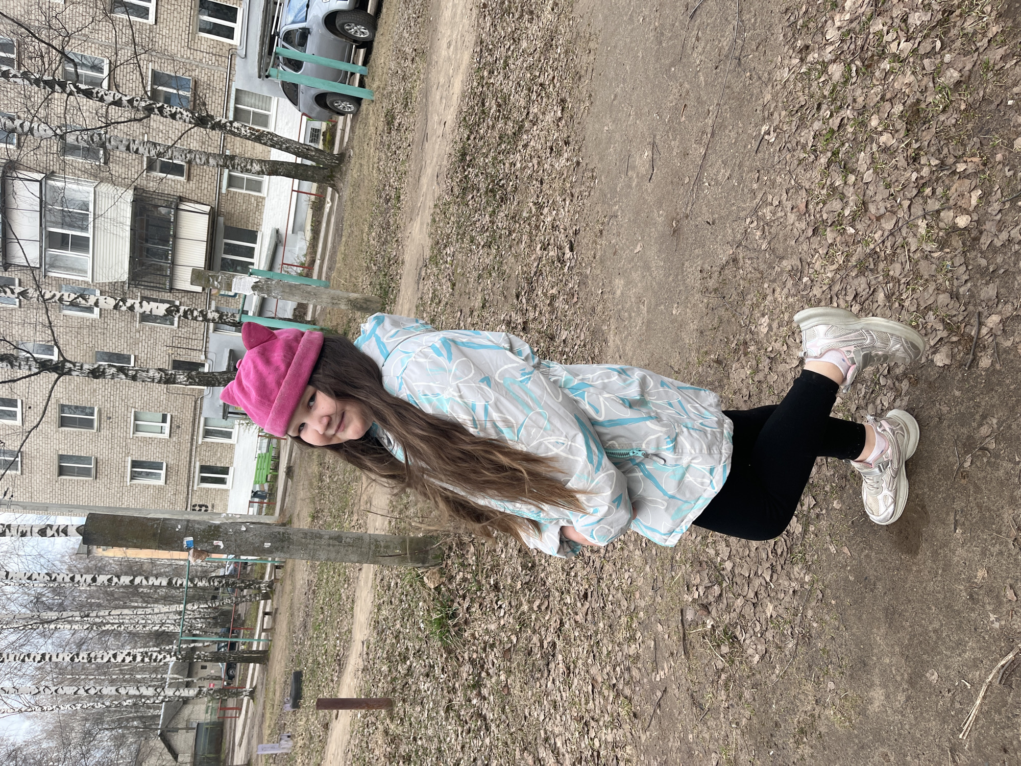 A young girl with long, dark brown hair stands outdoors in a residential area of Chaikovsky, Russia. She is wearing a pink beanie hat, a light blue and white patterned jacket, black leggings, and light-colored athletic shoes with pink socks. She looks towards the camera with a slight, pleasant smile.
The setting appears to be a paved area or unkempt ground covered with dry, fallen leaves and dirt, indicating late autumn, winter, or early spring. Several bare birch trees are scattered throughout the background, their white and black trunks prominent. Behind the trees, a multi-story, light-colored brick apartment building with numerous windows and balconies is visible. A grey car is parked near the building, and a wooden pole or post stands between the girl and the distant structures. The daylight suggests it is daytime, and the girl's attire implies cool or cold weather. No discernible text is visible in the image. The girl is simply posing for the camera.