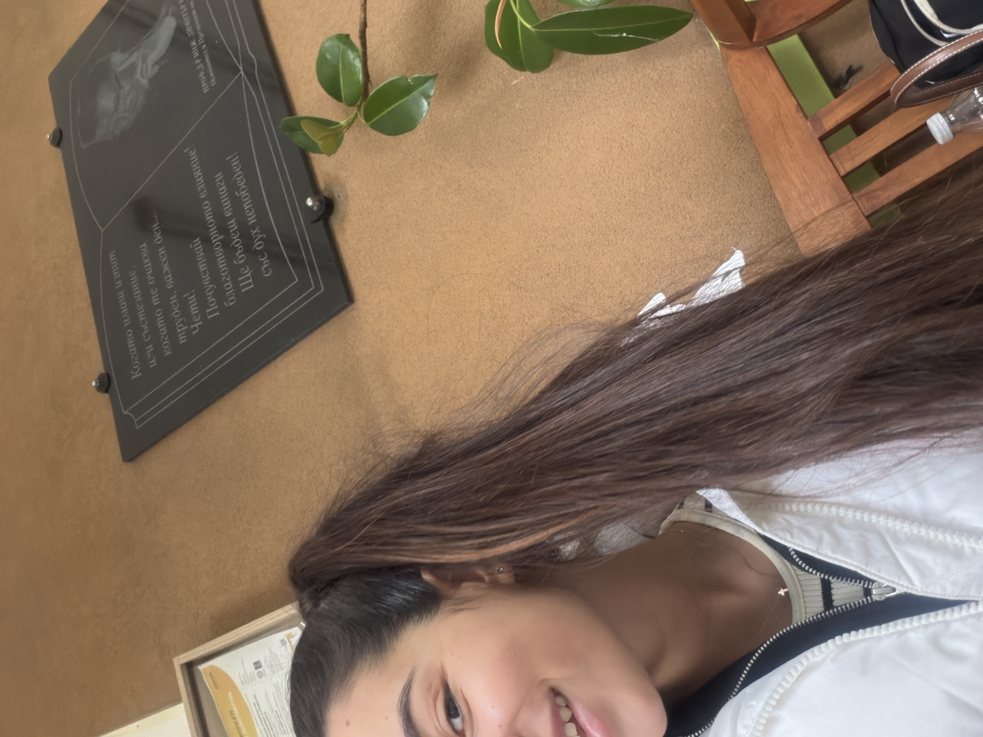 The image shows a person, likely a young woman, taking a selfie or photo from a close-up angle. She has brown hair and is wearing a white jacket. In the background, there is a brown wall with a black plaque attached to it, which appears to have some text in Bulgarian and a portrait. The text mentions "Когато имаш изпит" (When you have an exam) and "ПРОФ. Д-Р ИНЖ. ДИМИТРE" (PROF. DR. ENG. DIMITRE). There are also green leaves from a plant visible near the top of the frame. Part of a wooden chair with a bag on it is visible on the top right corner. The location appears to be indoors, possibly in a university building or historical place in Plovdiv, Bulgaria, as the Bulgarian text on the plaque suggests.
