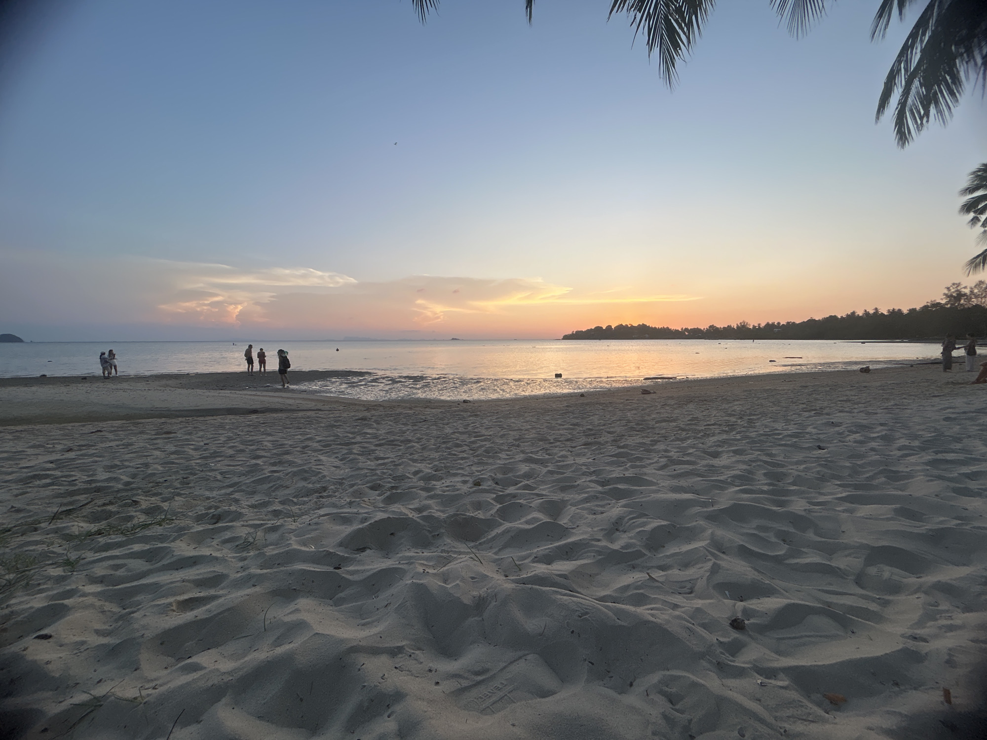 The image shows a beach at sunset, likely in Baan Sri Thanu, Thailand. The foreground is dominated by sand with visible footprints. In the middle ground, several people are standing at the water's edge. The sea is calm, reflecting the orange and pink hues of the setting sun. The horizon features a distant island or landmass. Palm trees frame the top of the image, adding to the tropical feel of the location.
