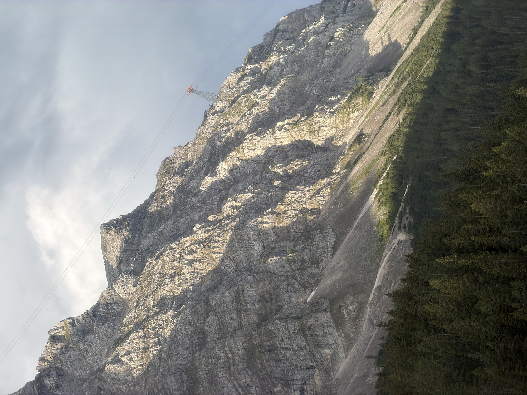 The image shows a steep, rocky mountain slope in Grainau, Germany. The mountain is covered in scree and sparse vegetation, with the sun casting dramatic shadows across its textured surface. A thick, dark green forest of coniferous trees covers the lower portion of the mountain. In the upper right portion of the image, a cable car pylon with a red car is visible, suspended by thin cables that stretch across the sky. The sky is partly cloudy, with a light gray hue. There are no people visible in the image.