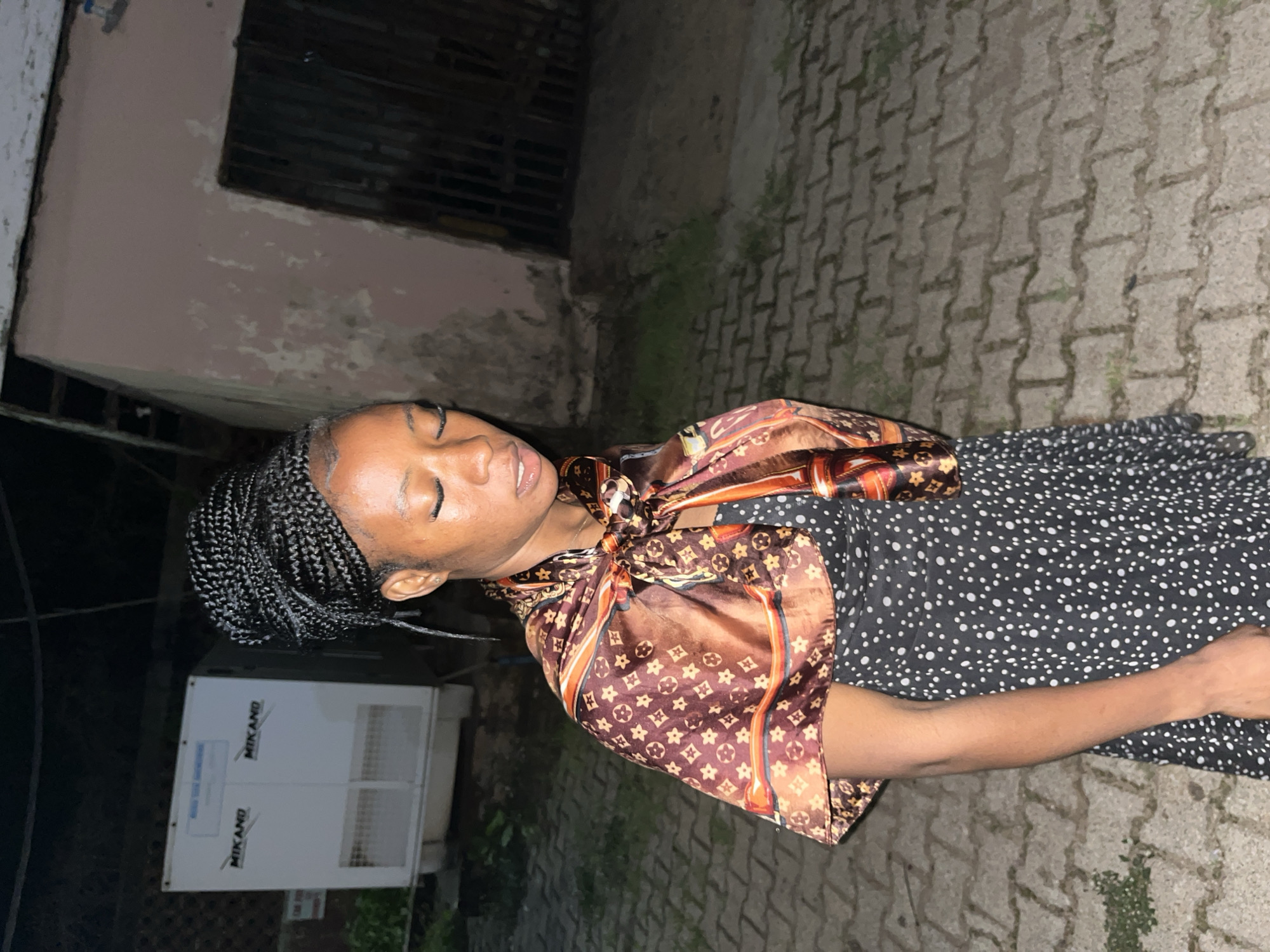 A young Black woman with braided hair is standing outside at night. She is wearing a black dress with white polka dots and a patterned scarf tied around her neck, draped over her shoulders. Her eyes are closed, and her head is slightly tilted down. The background shows a paved walkway, a wall of a building, and a large white appliance with "MIKANO" written on it. The setting appears to be an outdoor area in the city of Jos, Nigeria.