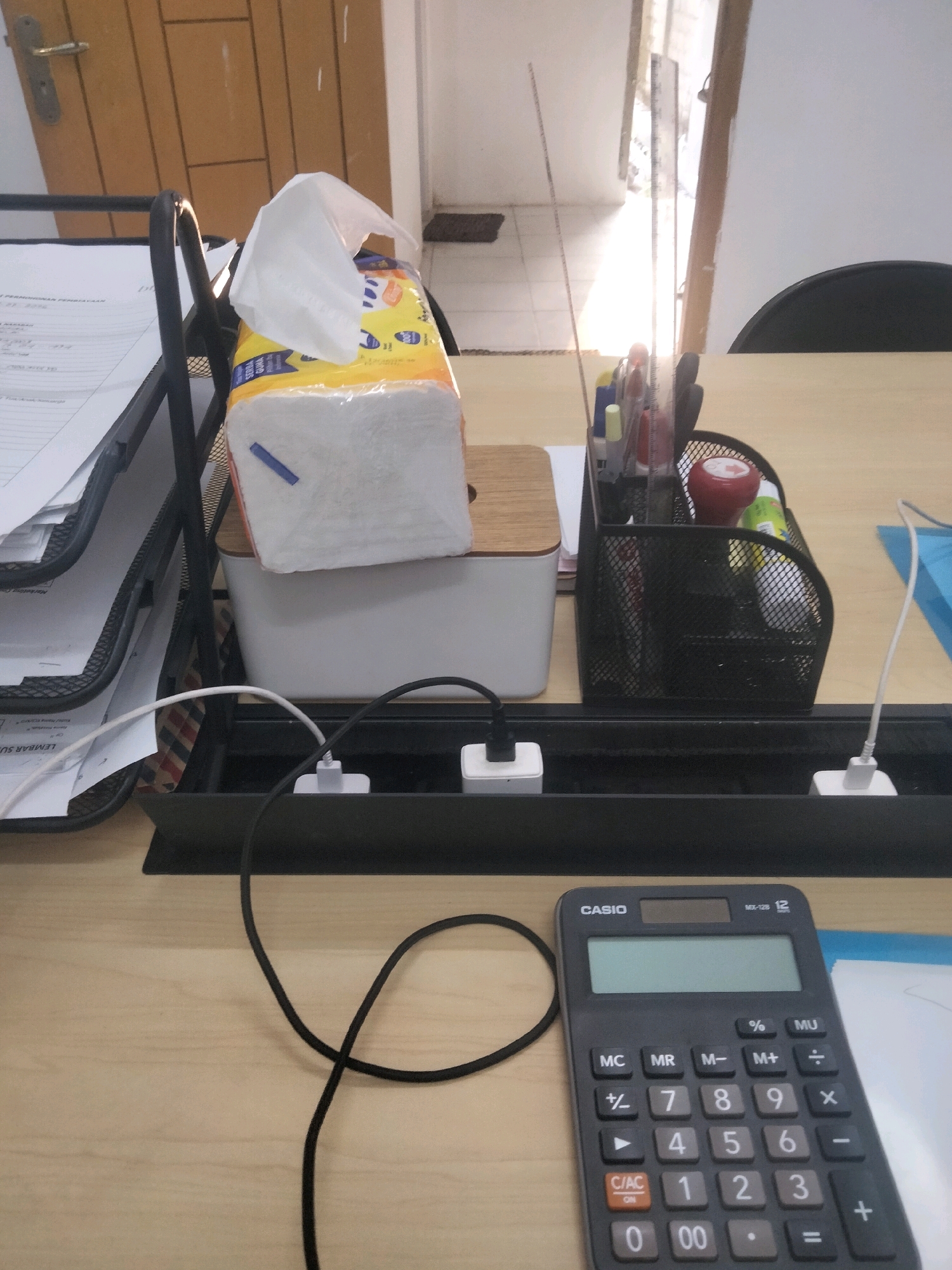 The image depicts a desk in an office in Seloretno, Indonesia. On the desk, a black mesh organizer holds pens and other office supplies. Adjacent to it, a white tissue box sits on top of a wooden and white container. In the foreground, a black calculator with a digital display is placed on the wooden surface. Various cables and power adapters are visible in a black cable management tray behind the calculator. To the left, a tiered document holder contains papers. The background shows a doorway with sunlight streaming in, illuminating a tiled floor.