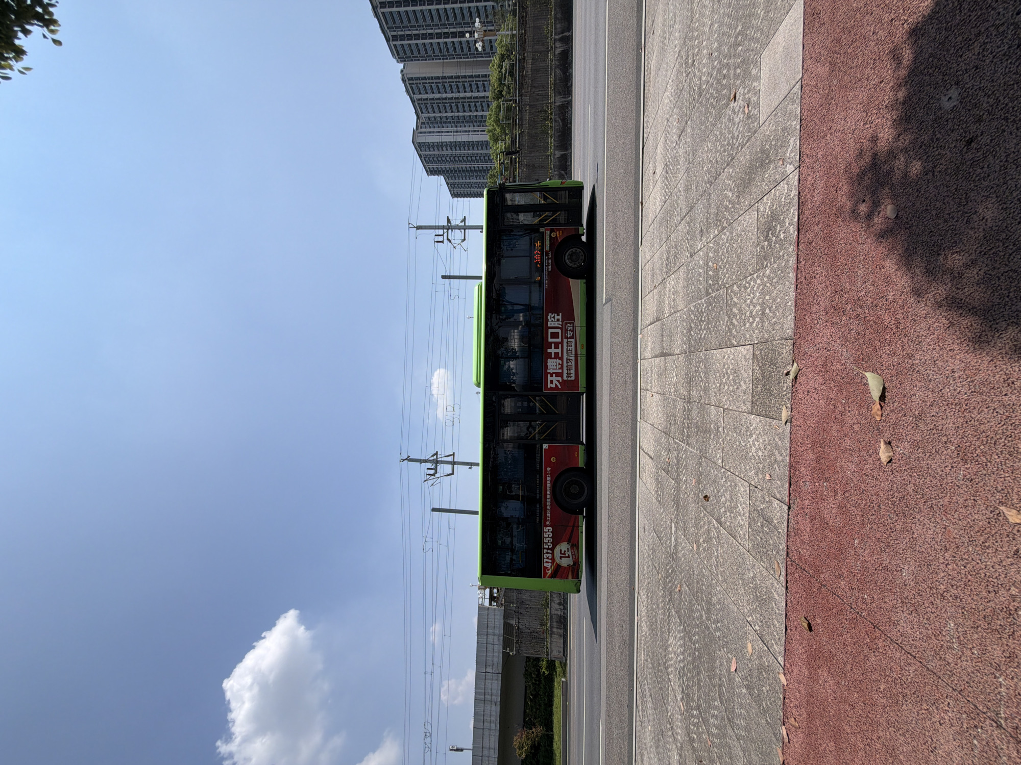 The image depicts a green and red bus driving on a road in Jiangjin, China. The bus is branded with Chinese text, including what appears to be a phone number "47375555" and the words "牙博士口腔" (Yá bóshì kǒuqiāng), which translates to "Dr. Dental Oral Cavity." The bus has advertisements on its side and displays "15" on a circular red emblem. In the background, tall apartment buildings line the horizon, with power lines and utility poles visible above a concrete barrier. The sky is clear blue with a single white cloud. The foreground shows a textured red and gray paved walkway with scattered fallen leaves. The scene is captured from a side-on, slightly low-angle perspective, suggesting the photographer is at ground level, possibly on the sidewalk. The overall impression is an urban street scene on a bright, sunny day.